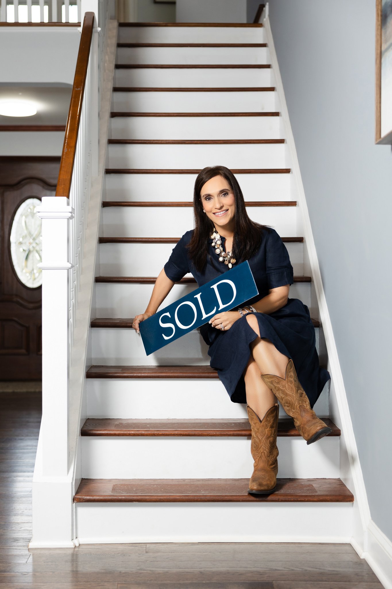 Woman sitting on staircase holding a "SOLD" sign, smiling, inside a house with wooden floors and a front door in the background.
