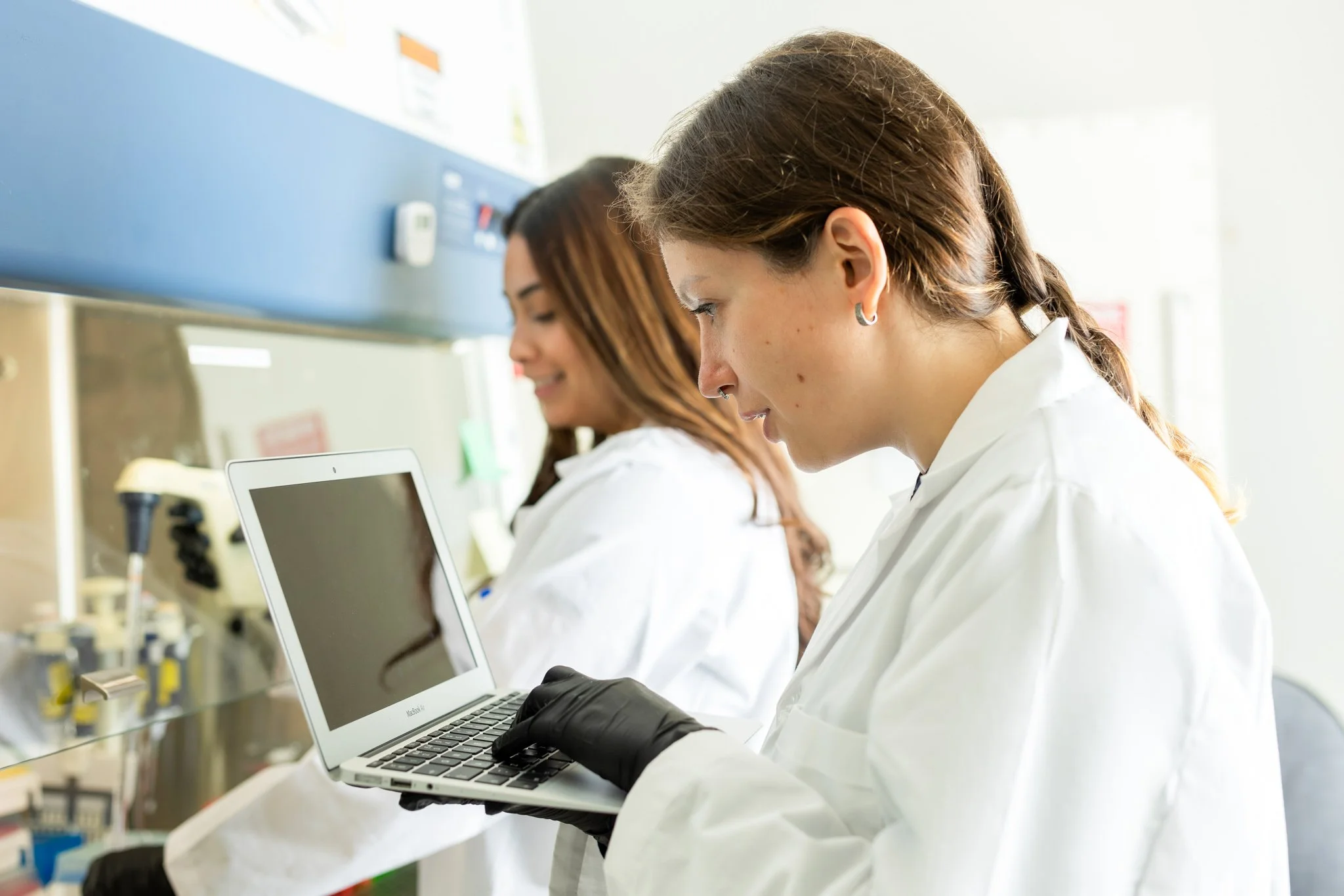 Two female scientists in white lab coats working in a laboratory, one using a small laptop, with lab equipment in the background.