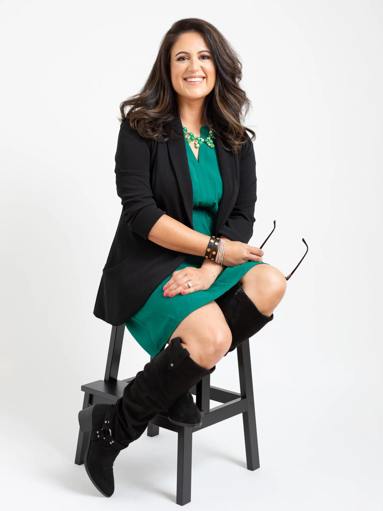A woman with long, wavy brown hair, wearing a teal dress, black blazer, black knee-high suede boots, and a green statement necklace, sitting on a black chair against a white background, smiling, holding glasses in her right hand.