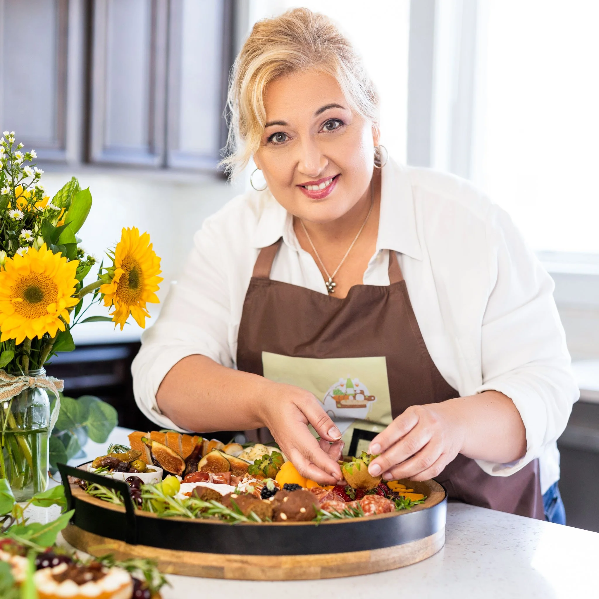 A woman in a white shirt and brown apron arranging food on a round platter in a kitchen, with a bouquet of sunflowers and a cupcake nearby.