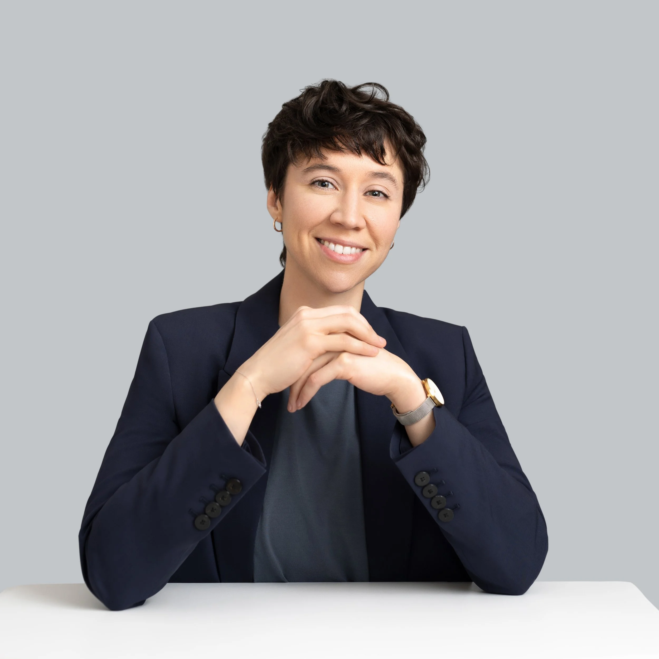 Smiling woman with short brown hair wearing a dark blazer and gray shirt, sitting at a white table against a plain gray background.