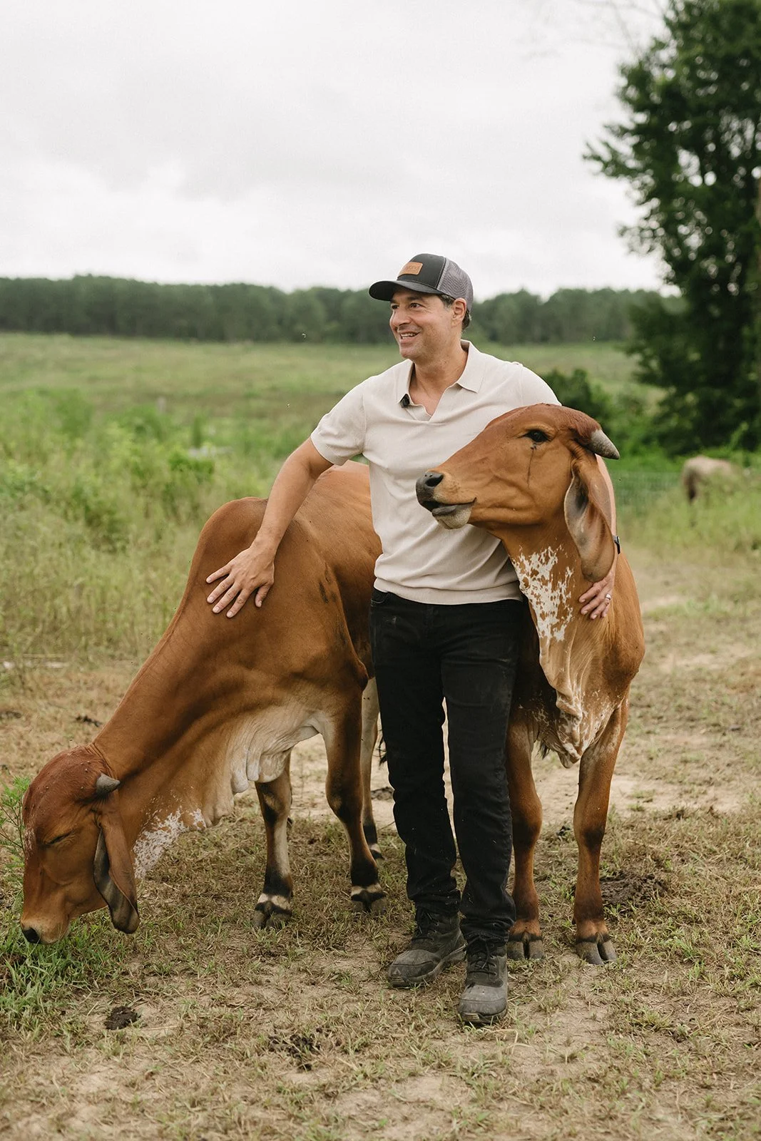A man in a white shirt and black pants hugging two brown cows in a grassy field, smiling, with trees and an overcast sky in the background.