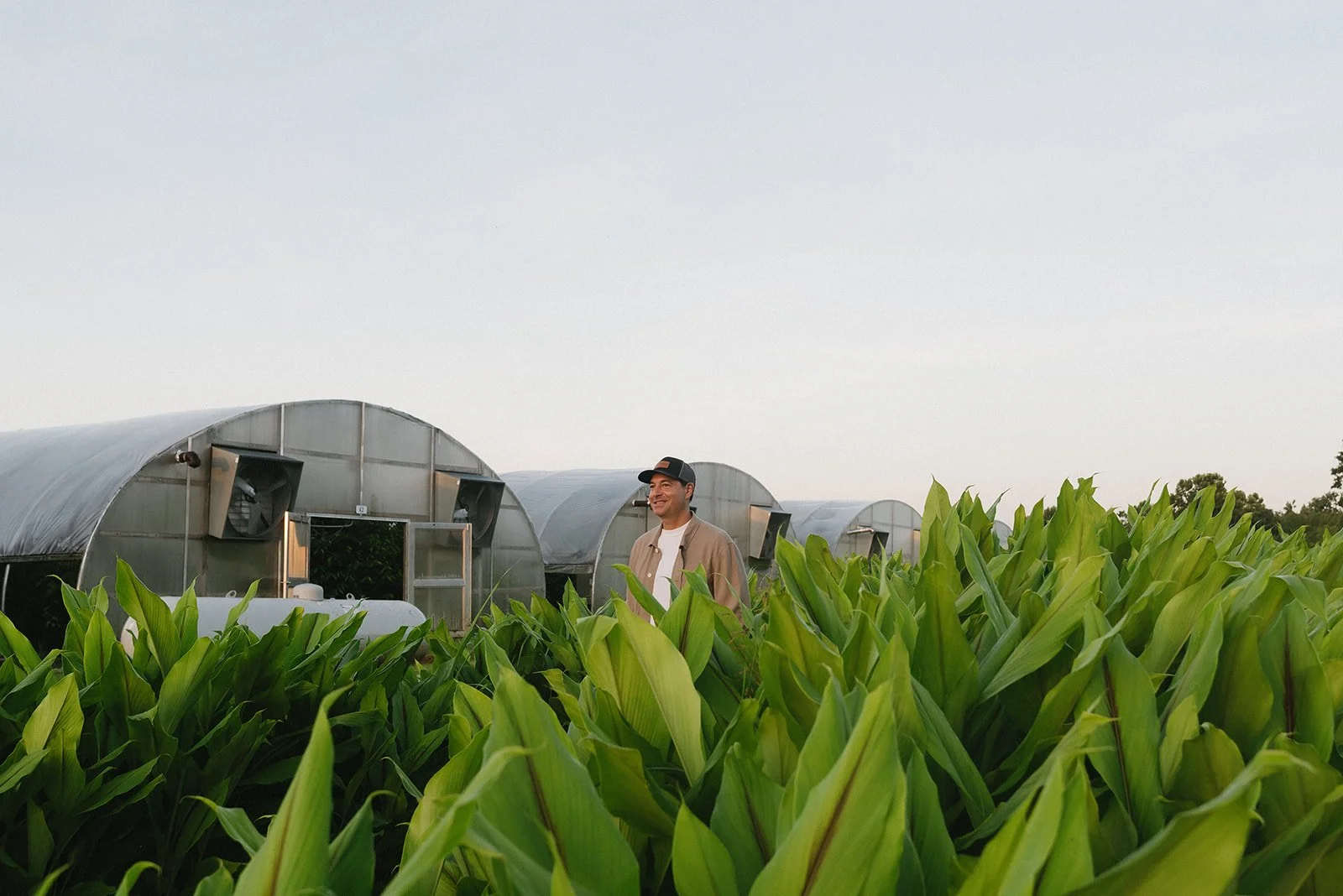 Man smiling in a green field with greenhouse structures in the background.