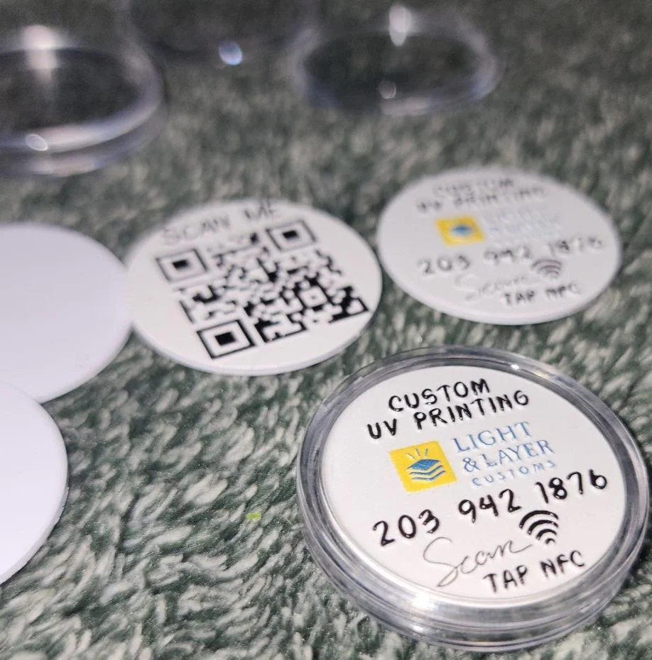 Collection of round adhesive labels on a textured surface, with one label reading 'CUSTOM UV PRINTING, LIGHT & LAYER, 203 942 1876, tap NFC' and others including QR codes and contact information.