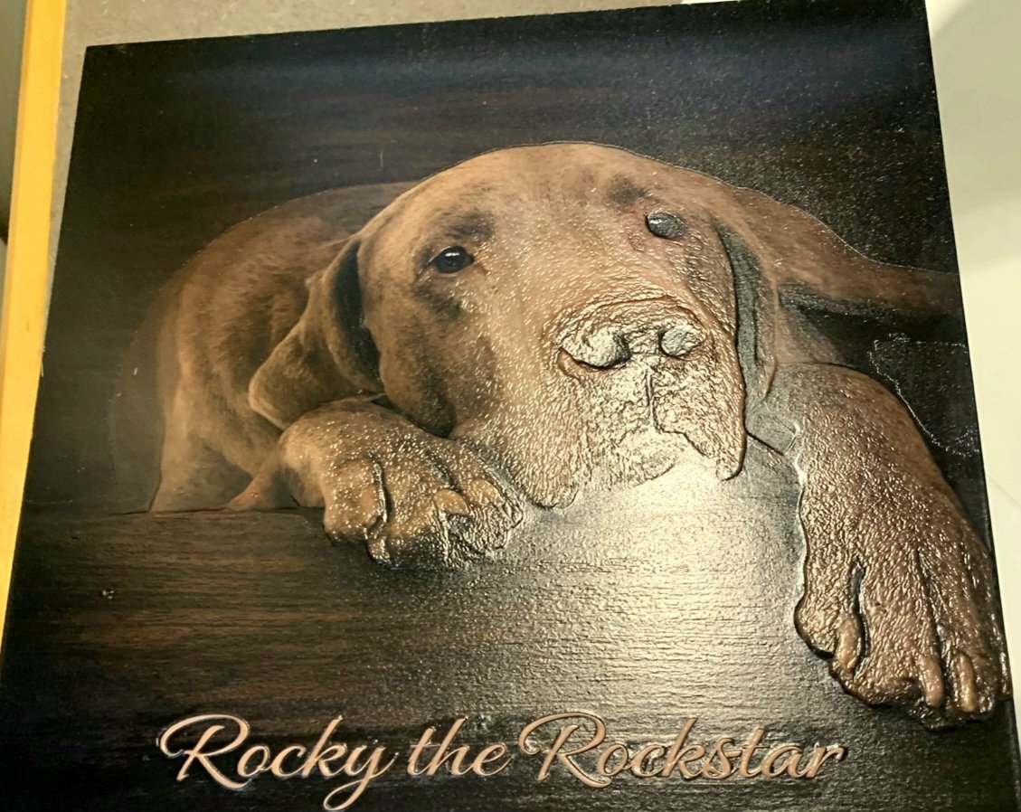 Wooden plaque with an engraved image of a resting brown dog with a happy expression, paw extended, and the text "Rocky the Rockstar" beneath.