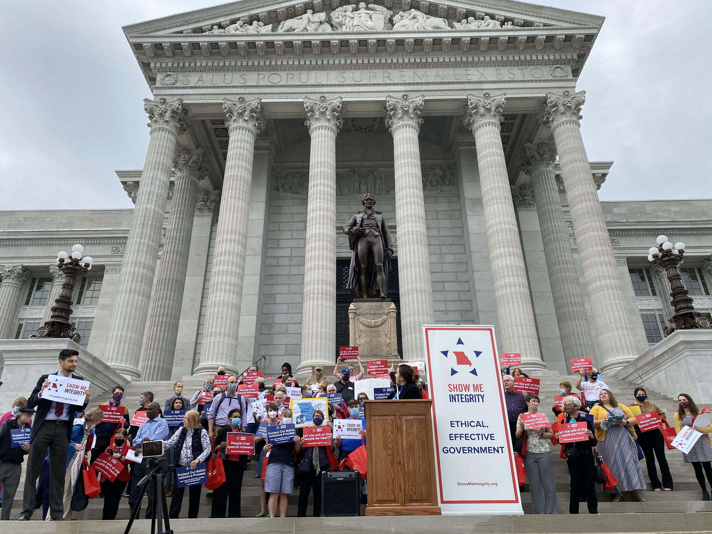 A group of people holding signs in front of the Missouri State Capitol Building. Several of the signs read "Show Me Integrity"