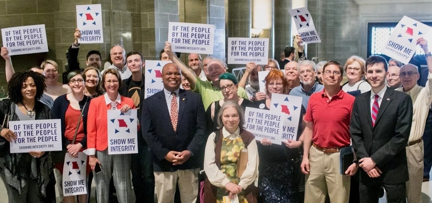 A group of people holding signs. Many of the signs read "Of the people, by the people, for the people."