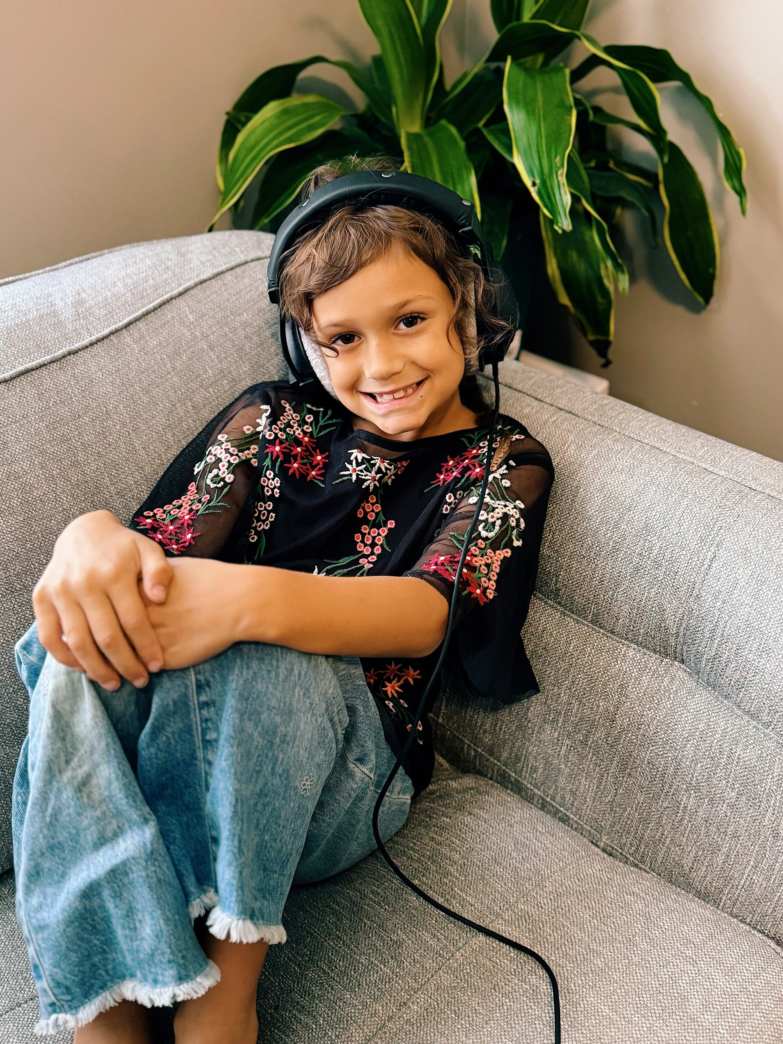 A young girl with curly brown hair and a smile, wearing black headphones and a black embroidered top, sitting on a gray sofa with her legs crossed, in front of a green houseplant.
