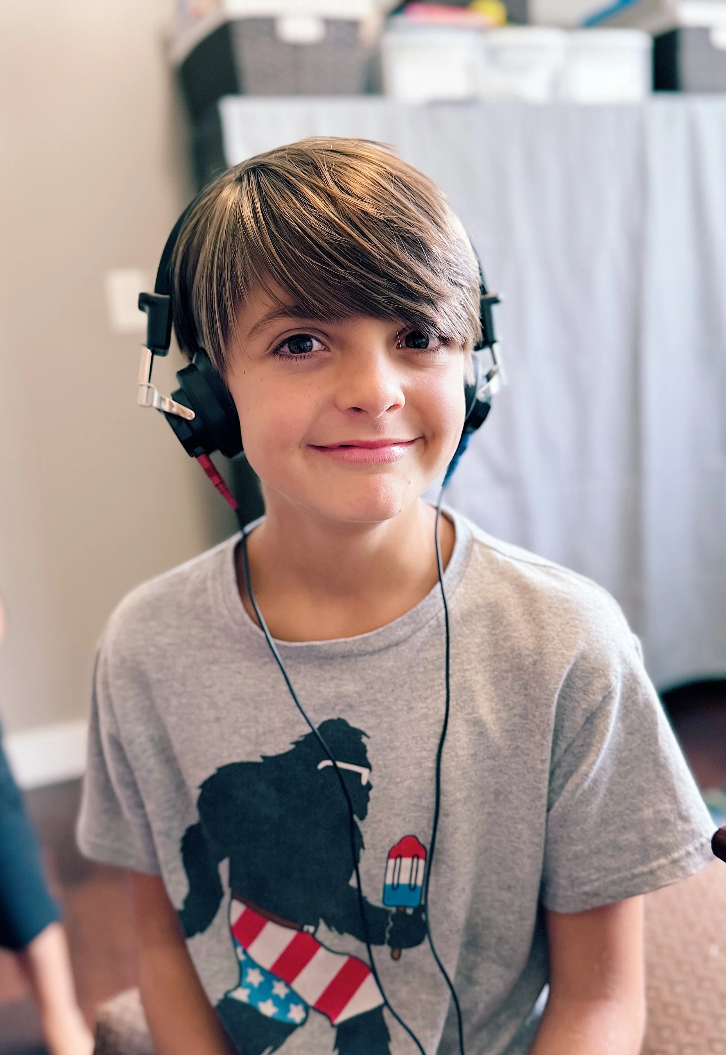 A smiling young boy with light brown hair wearing a gray T-shirt with a graphic of a French bulldog, American flag pattern, and a popsicle, and black over-ear headphones, indoors.