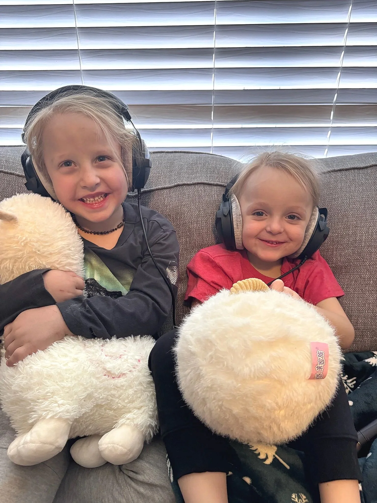 Two young children, a girl with a black shirt and a girl in a red shirt, sitting on a couch with plush stuffed animal toys, wearing headphones, with window blinds in the background.
