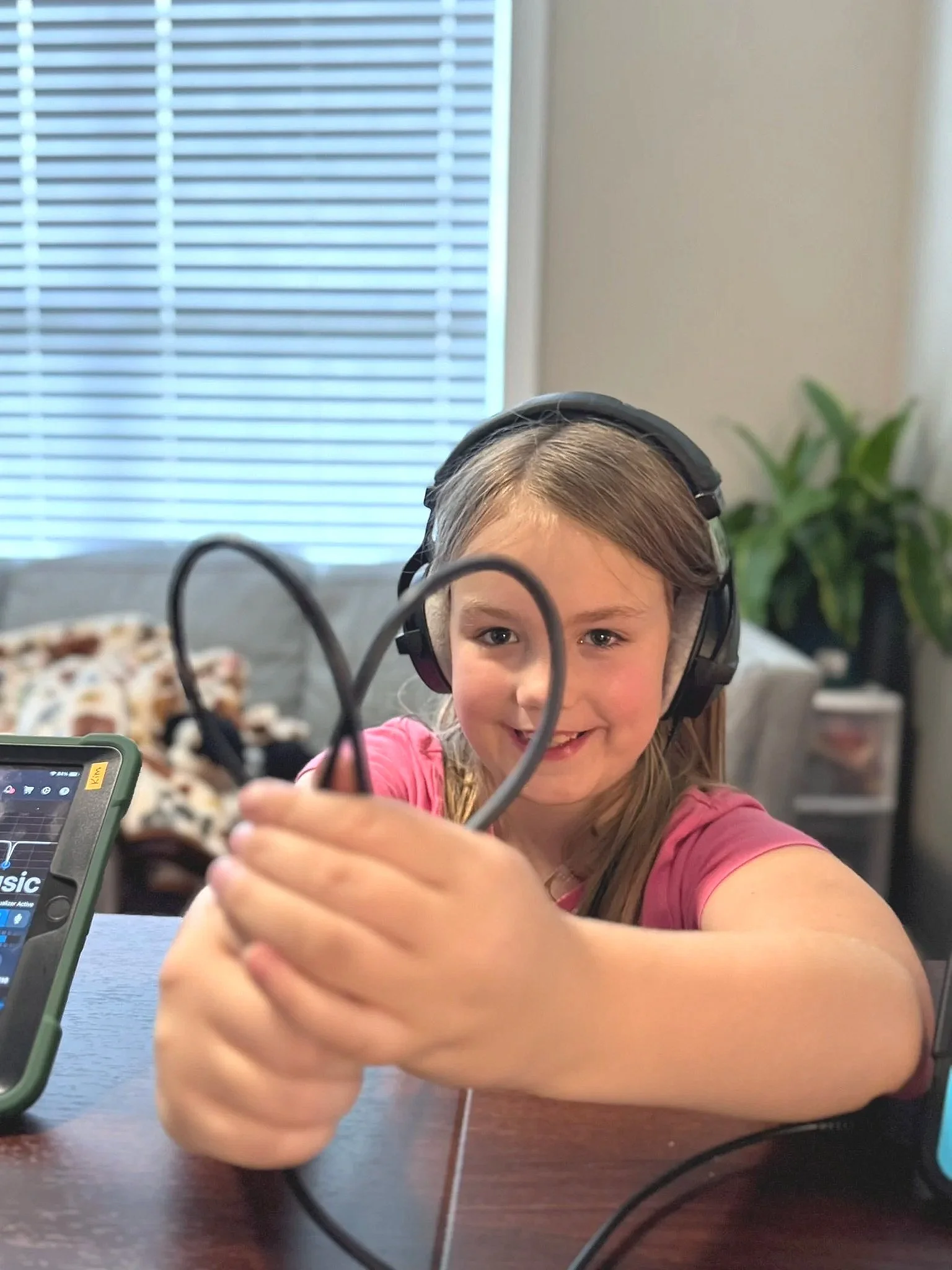 A young girl wearing headphones is holding audio cables and smiling at the camera, sitting at a table with electronic devices in a room with a window and a tall green plant in the background.