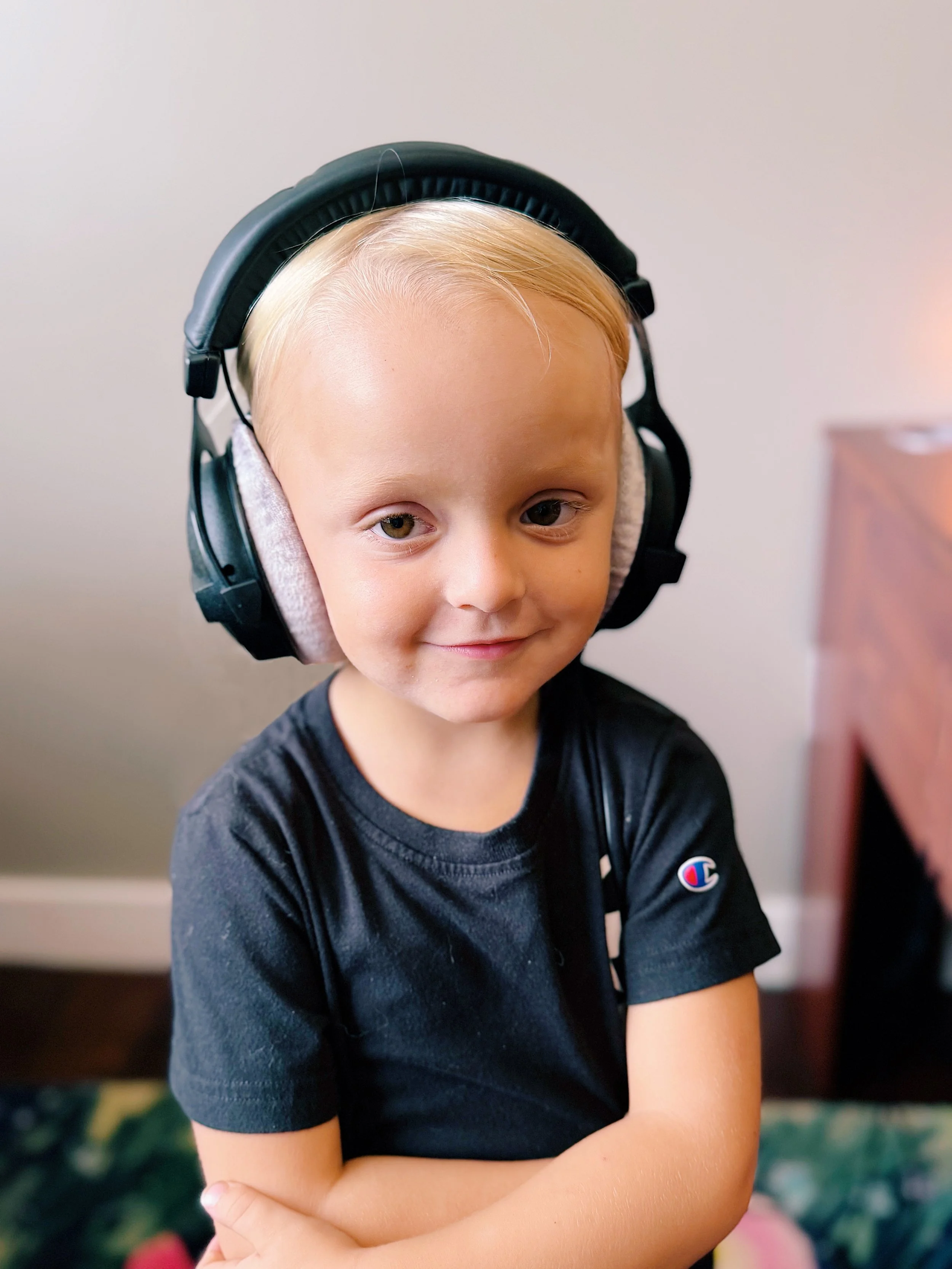 A young boy with blonde hair wearing a black t-shirt and large black headphones, smiling with arms crossed inside a room.