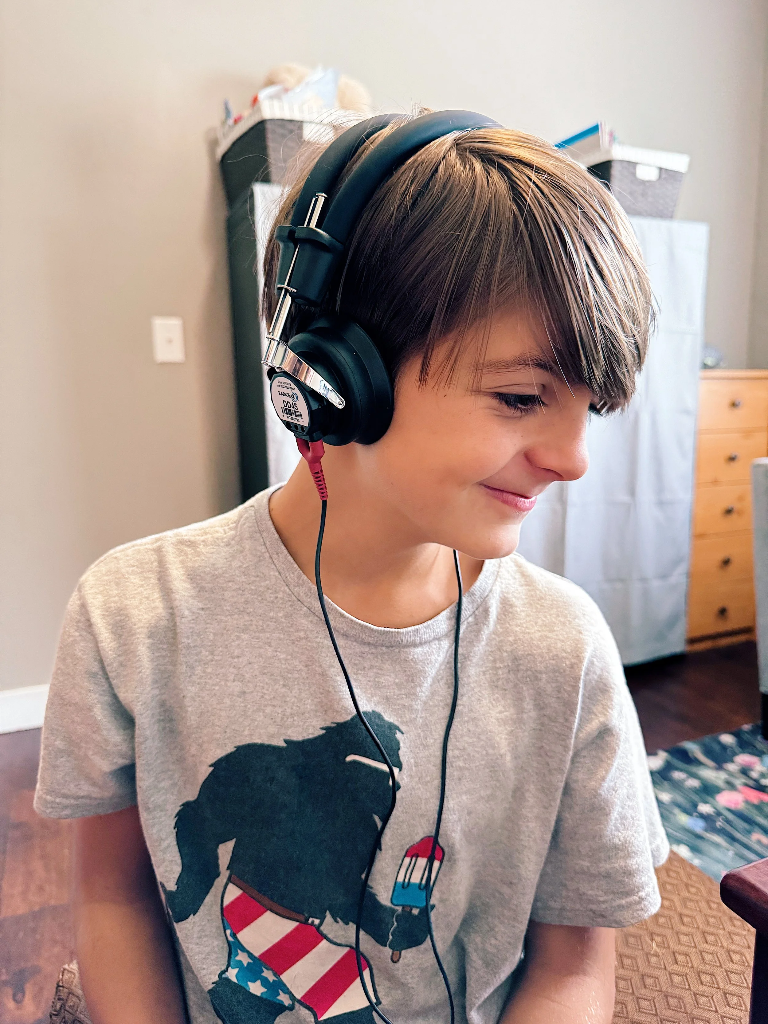 A young boy with brown hair wearing headphones and a gray T-shirt with a graphic of a dog holding a popsicle, smiling with his eyes closed in a room with furniture and a floral area rug.