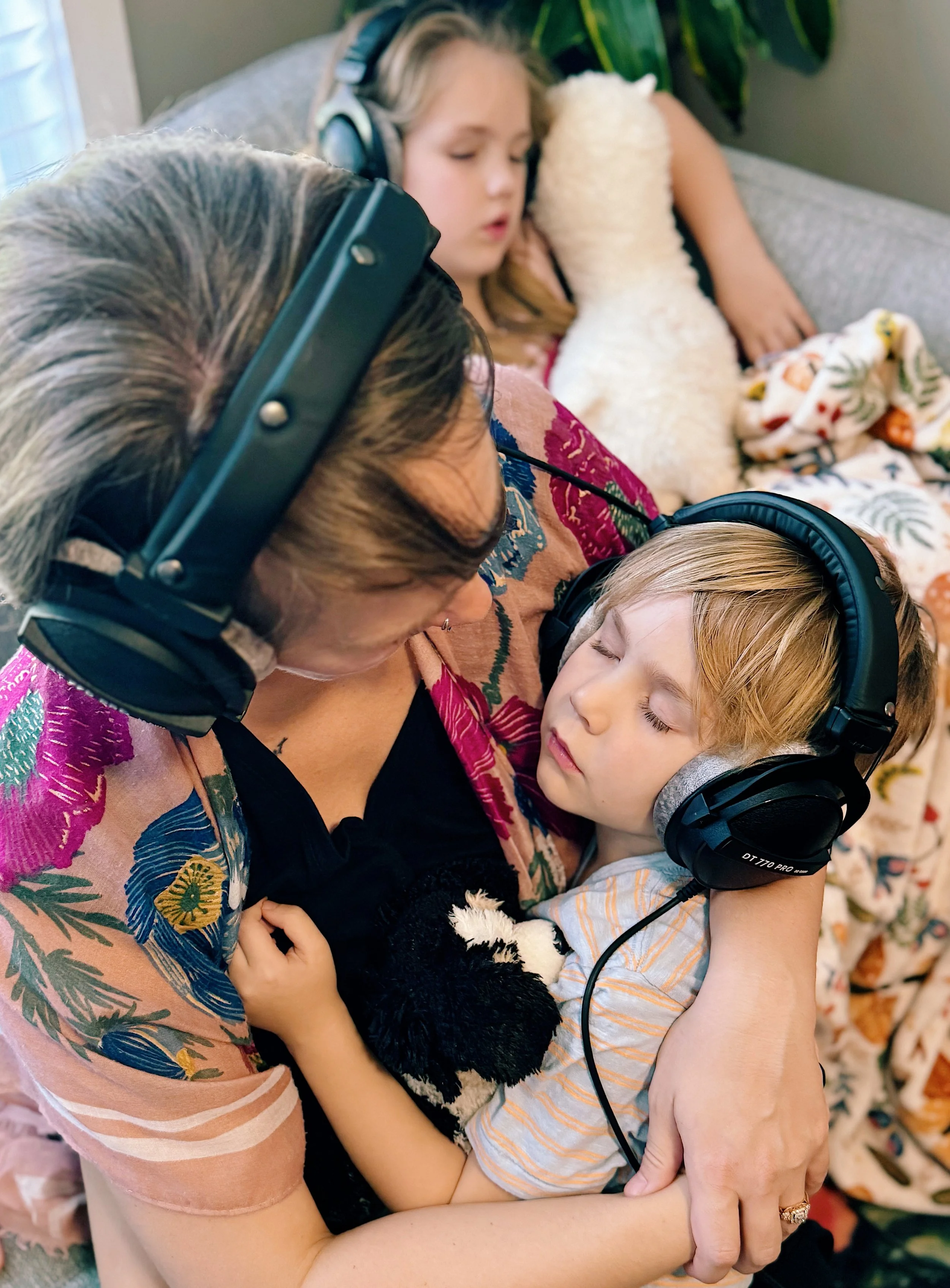 A woman and two children, all wearing headphones, sitting on a couch. The woman is holding one child in her arms, and the other child is in the background, sleeping with a stuffed animal. The scene appears cozy and intimate.