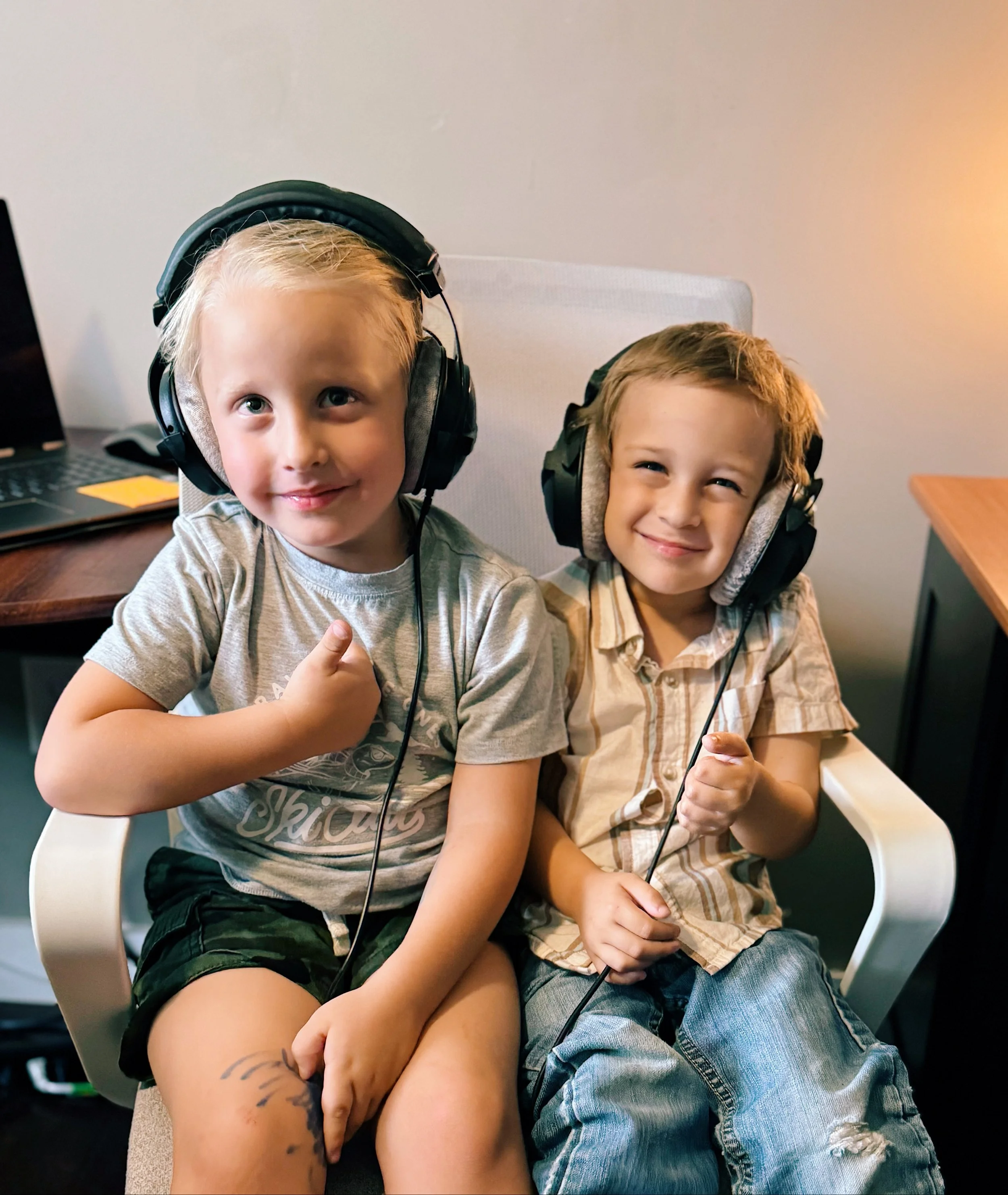Two young boys sitting on a chair wearing headphones, smiling at the camera, in a room with a light-colored wall and a desk nearby.