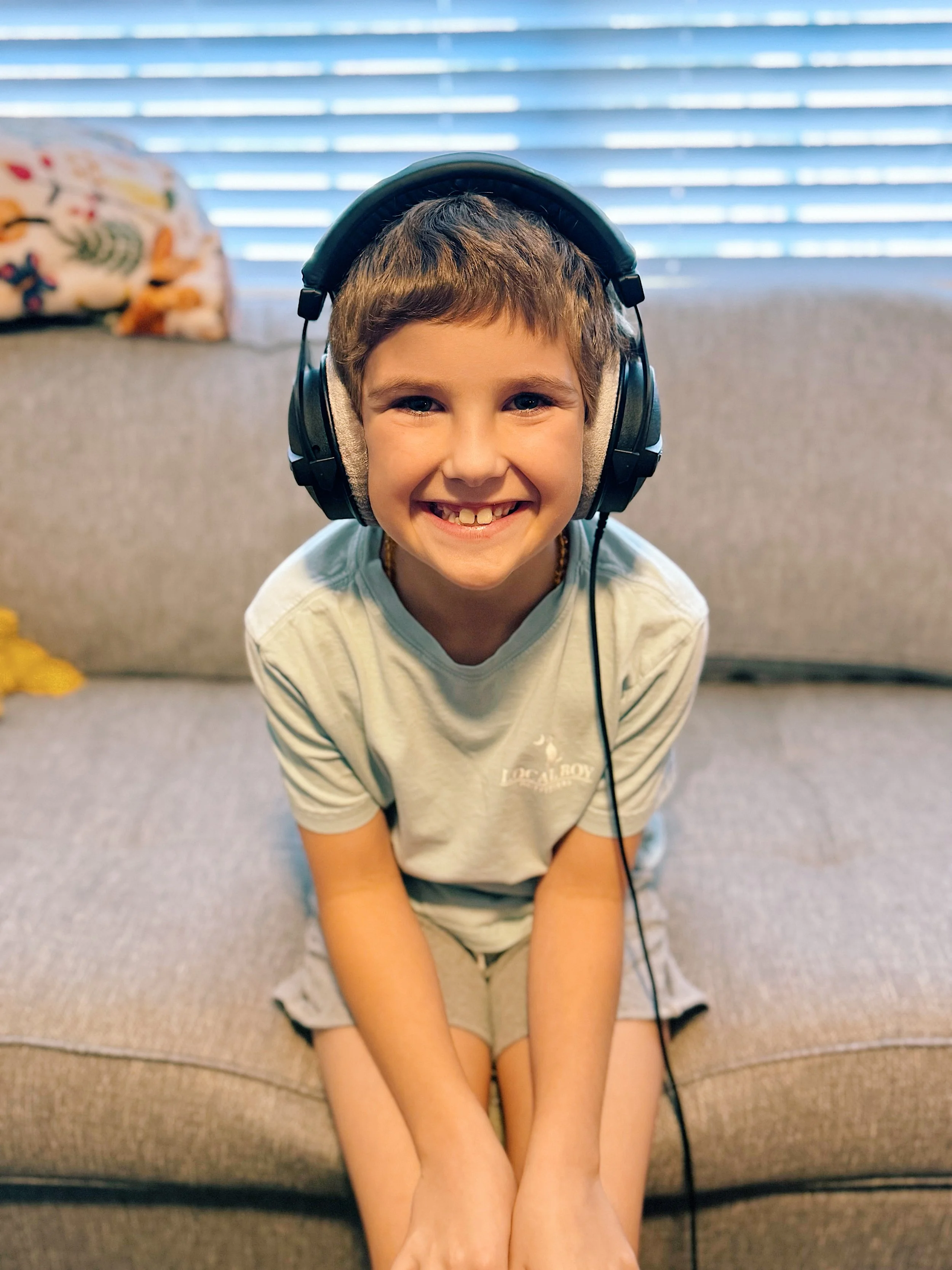 A smiling boy with brown hair wearing headphones, sitting on a gray couch with blinds in the background.