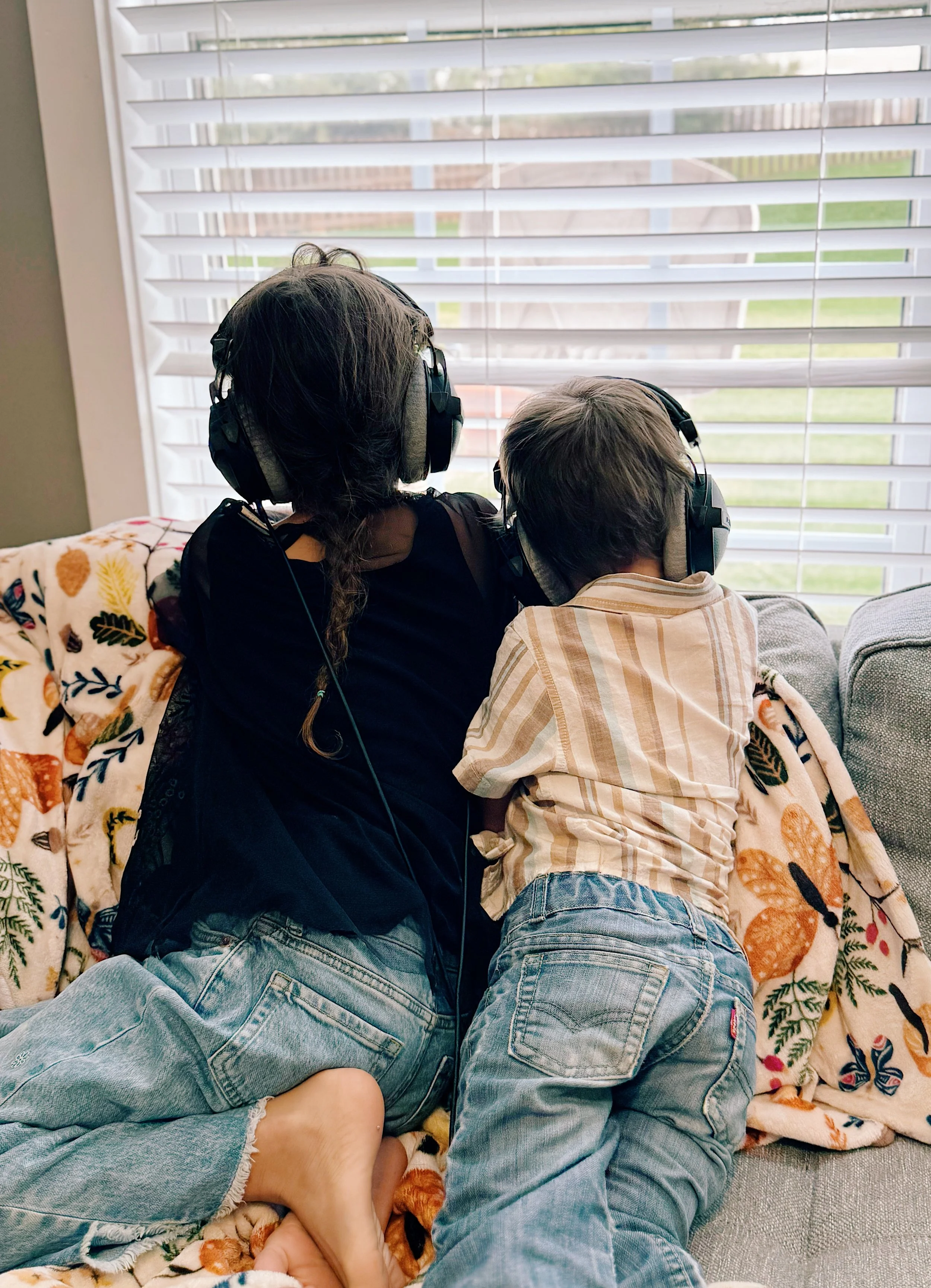 A woman and a young boy sitting on a couch, both wearing headphones, looking out the window with blinds. The woman is wearing a black top and faded jeans, while the boy is in a striped shirt and jeans. There is a floral blanket on the couch.
