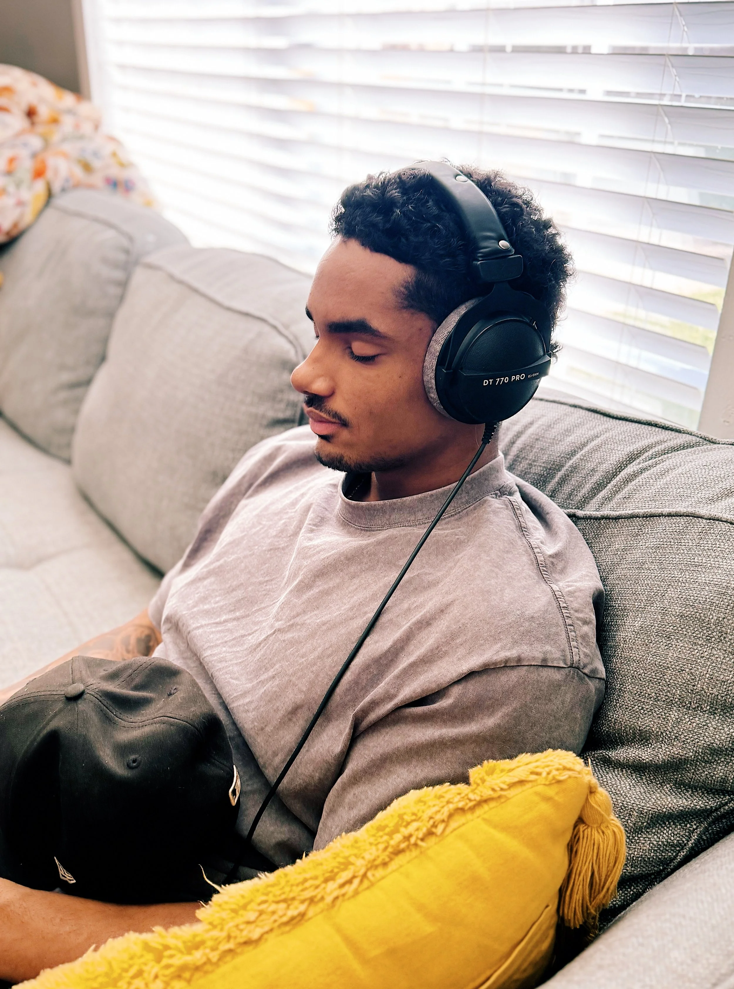 A young man with dark curly hair is sitting on a gray fabric sofa, wearing large over-ear black headphones with a gray headband. He has closed eyes and appears relaxed. There is a yellow pillow with tassels on his right side and a black baseball cap 