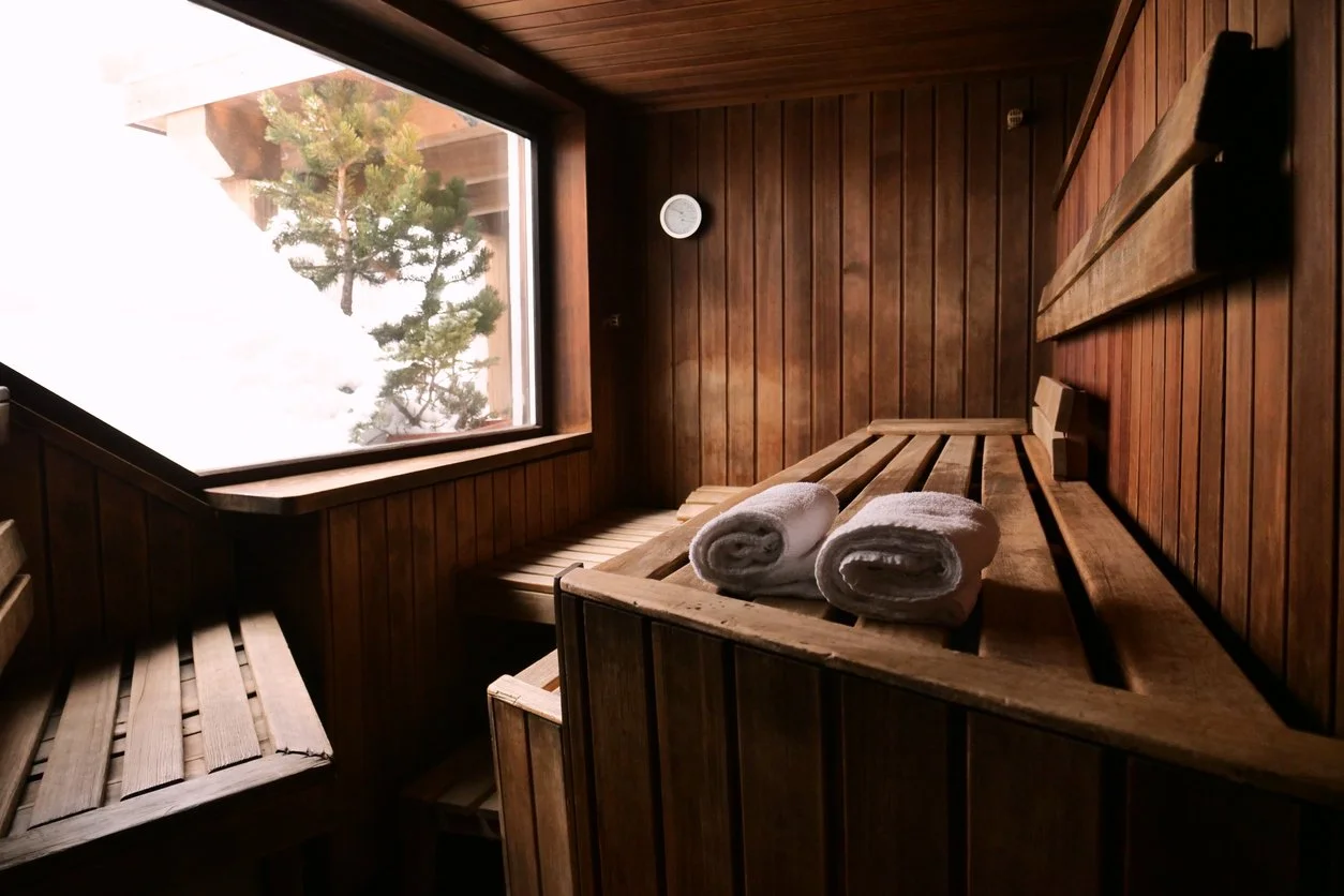 Wood-paneled sauna room with a large window showing snow and a pine tree outside, two rolled white towels on a wooden bench, and a wall-mounted thermometer.