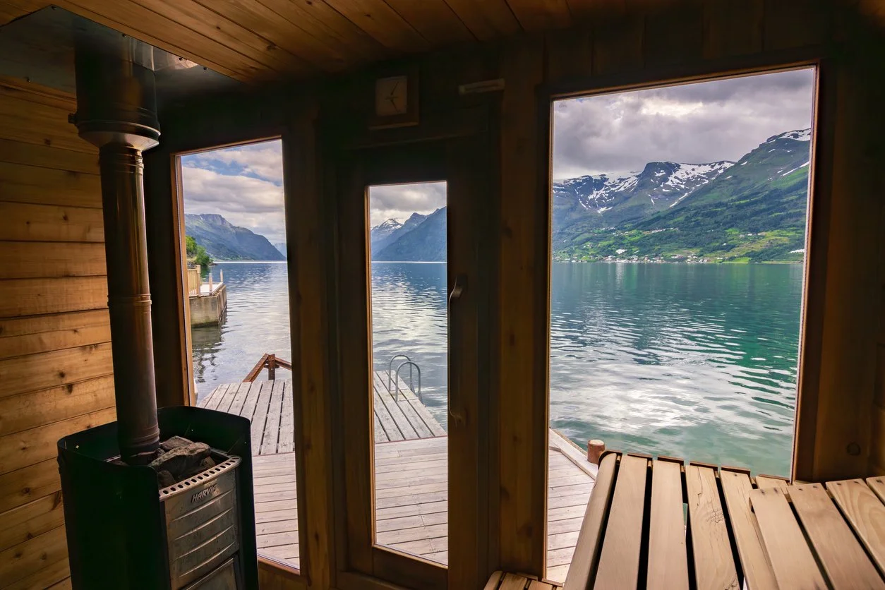 Interior view of a wooden sauna overlooking a scenic lake surrounded by mountains with snow-capped peaks.