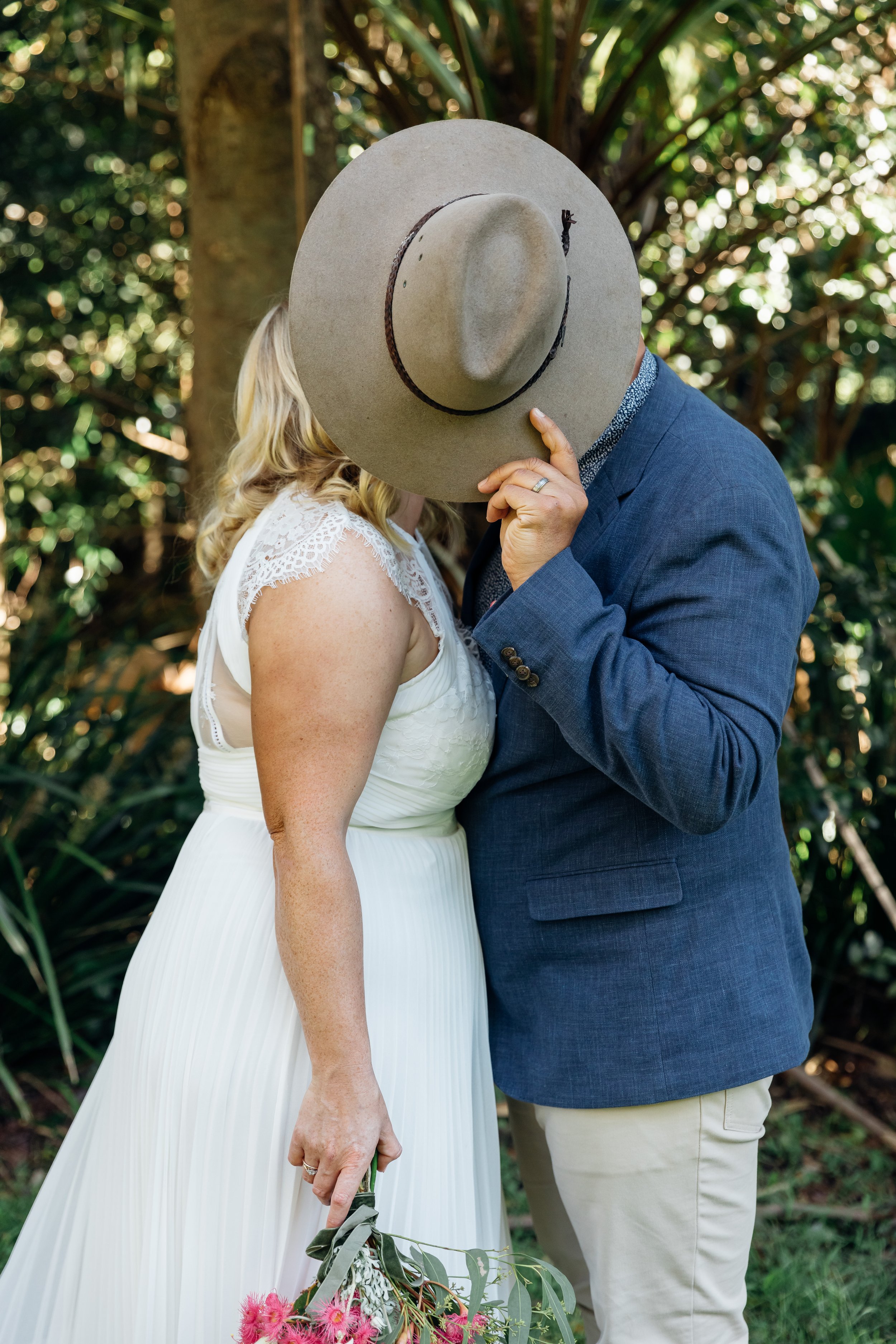 A couple dressed in wedding attire, with the woman holding a bouquet of pink flowers, sharing an intimate moment outdoors while the man covers their faces with a large hat in a lush, green setting.