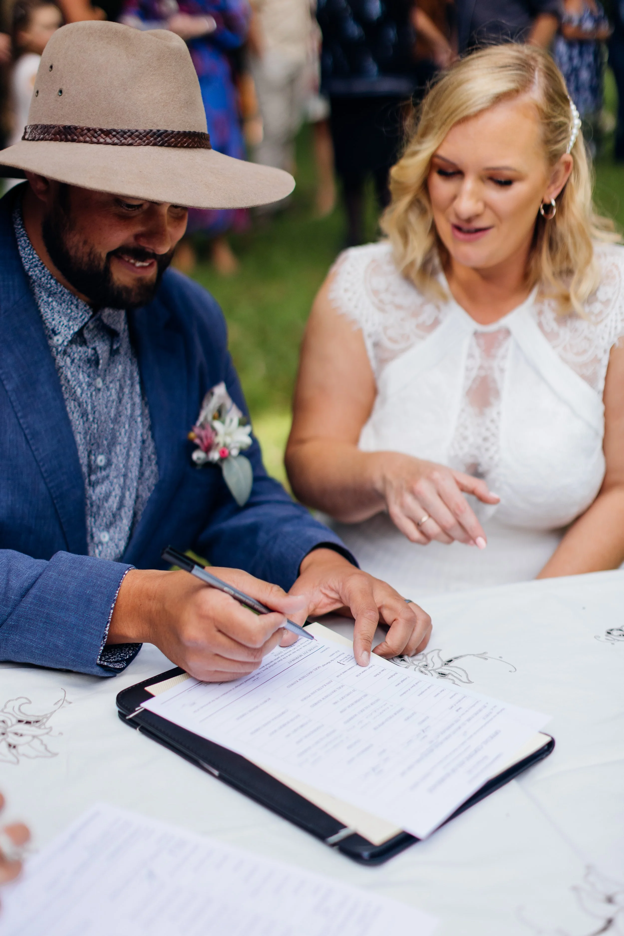 A man in a wide-brimmed hat and a woman in a white lace dress signing a document at a wedding ceremony.