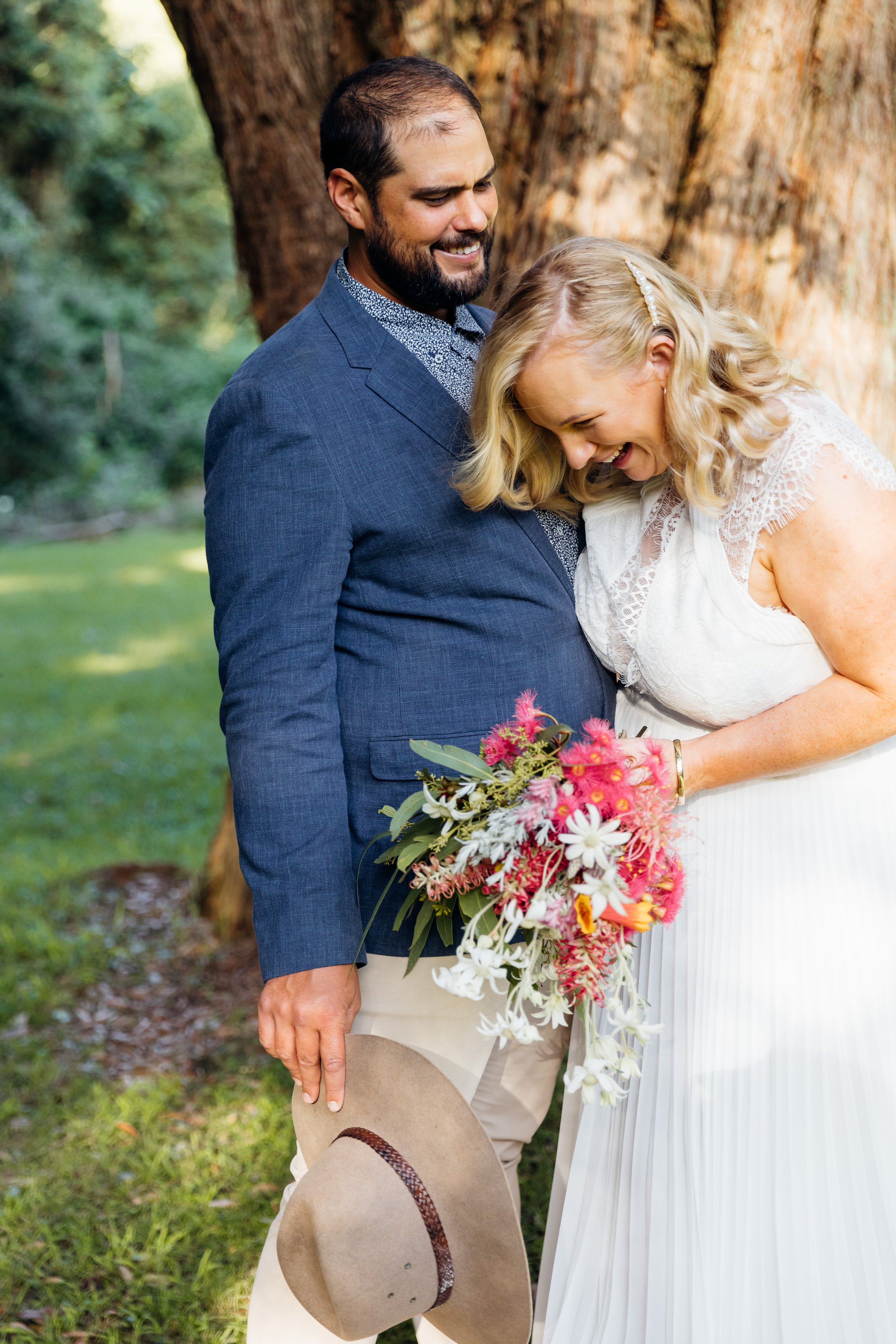 A bride and groom sharing a joyful moment outdoors near a large tree, with the bride holding a colorful bouquet and the groom holding a beige hat.