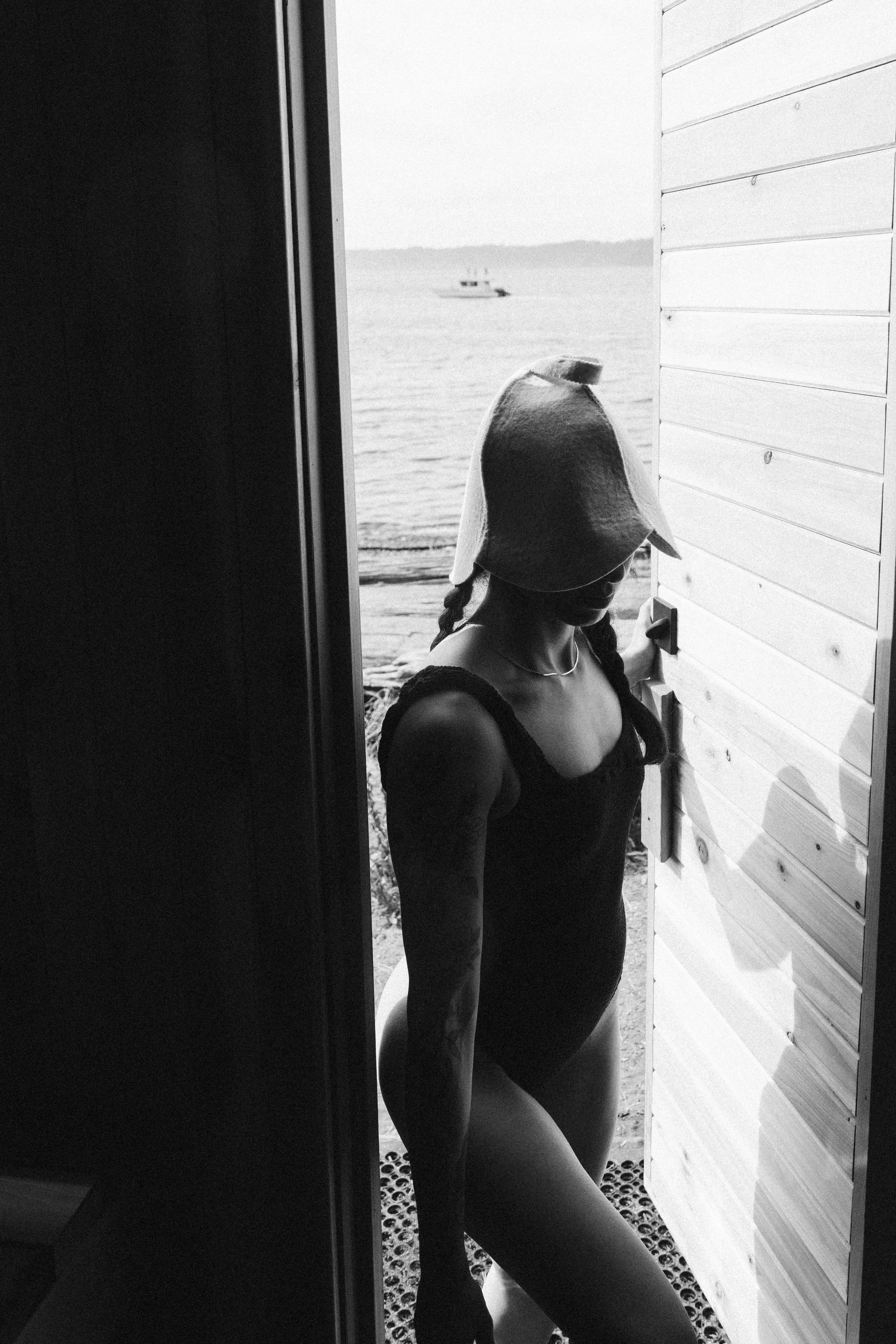 A girl with braided hair, wearing a hat and a sleeveless top, standing on a porch with her hand on the door handle, looking at a boat on the water in the distance.