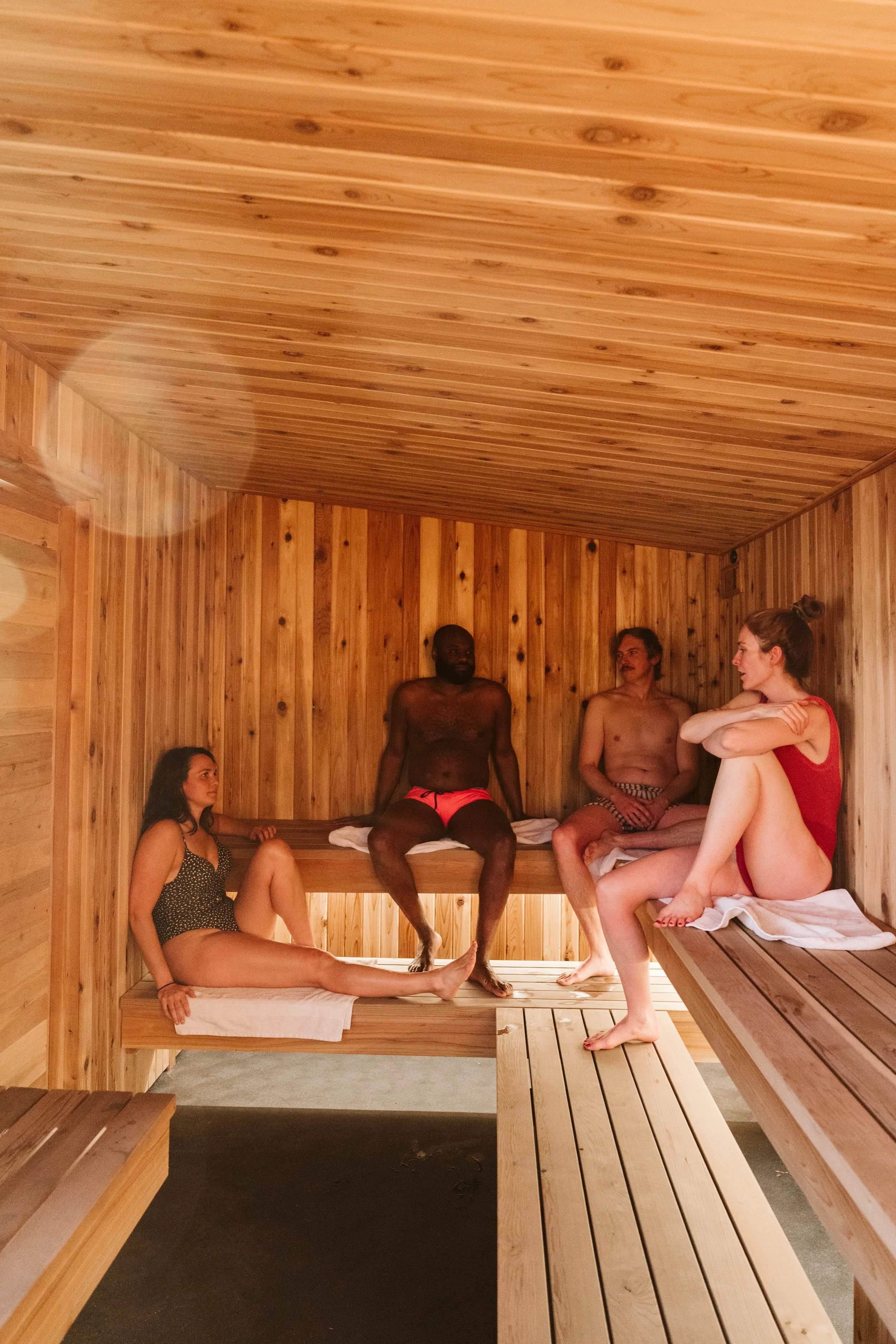 Several people inside a small wooden sauna, sitting or relaxing on benches, with wood-paneled walls and ceiling.