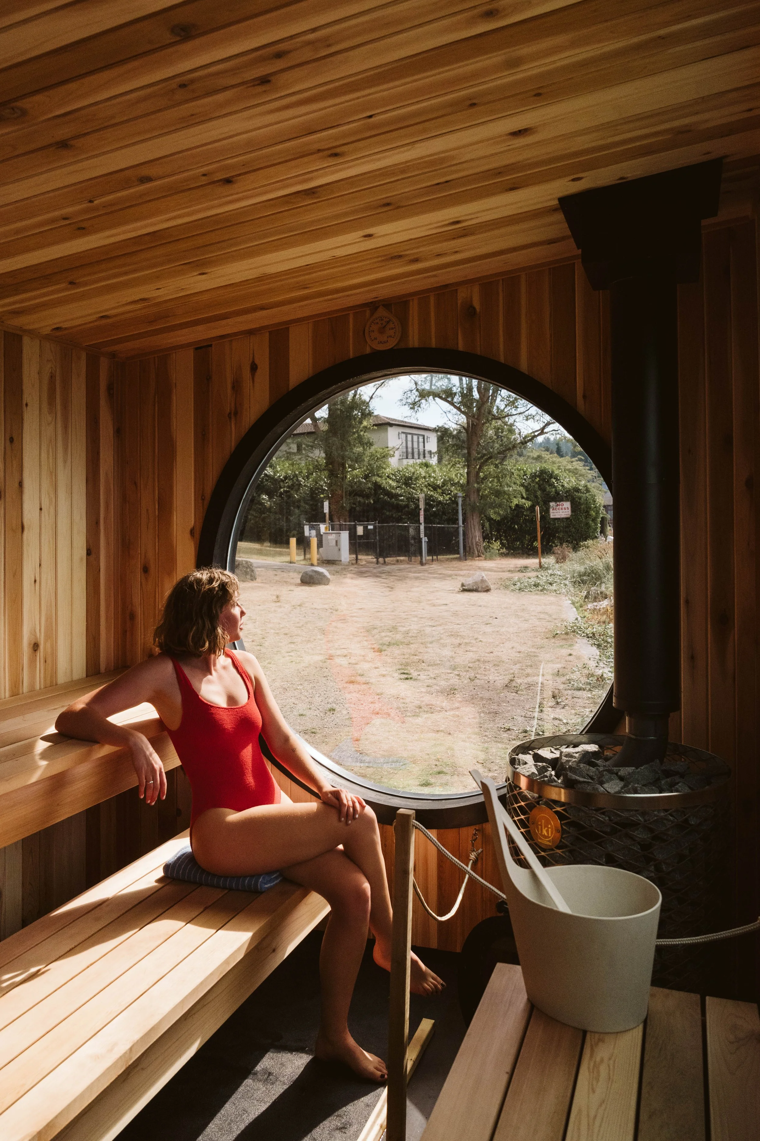 A woman in a red swimsuit sitting inside a wooden sauna overlooking an outdoor scene with trees and buildings through a circular window. There is a stove with rocks in the corner and a thermometer on the wall.