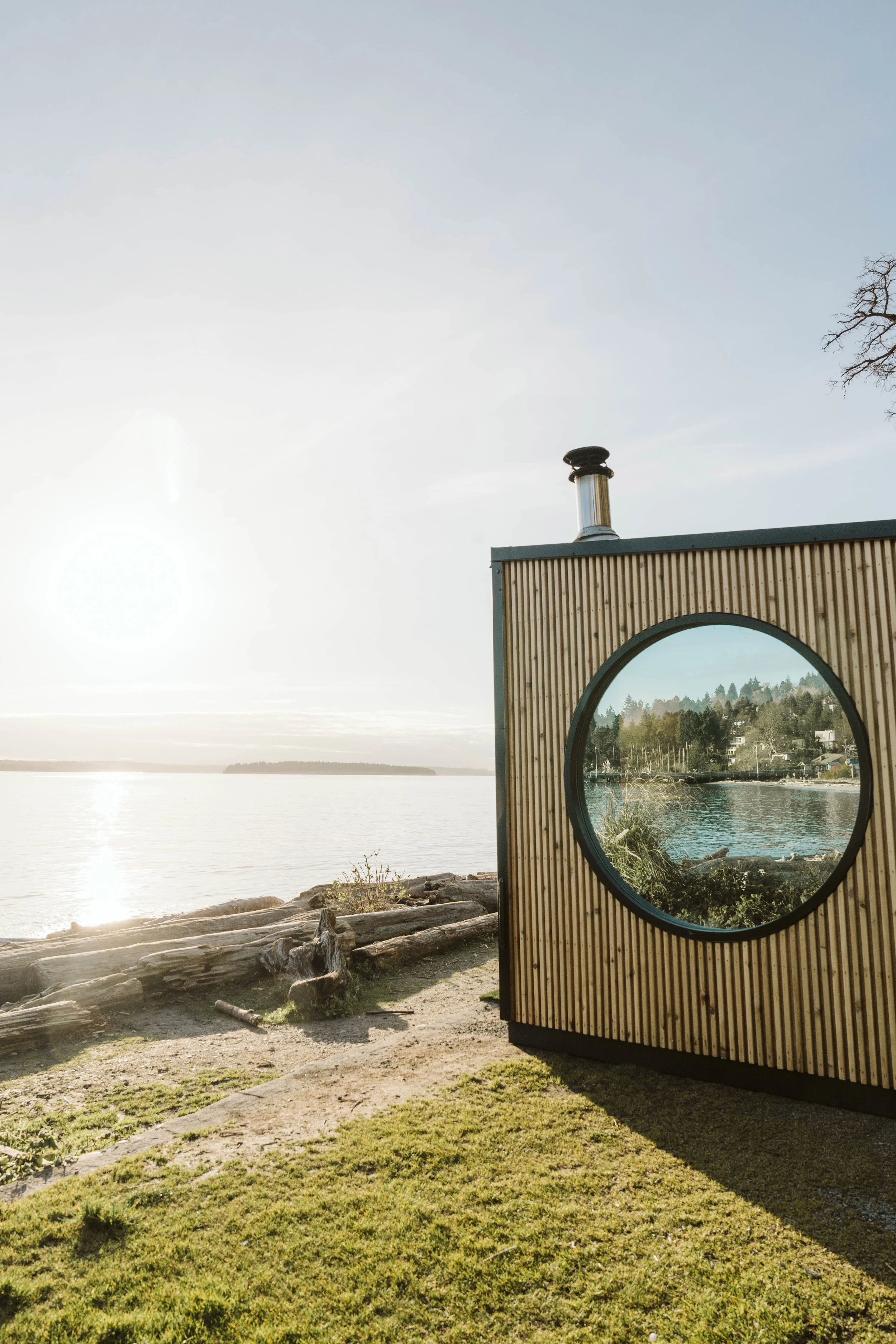 Sauna building with a circular window, reflecting trees, situated near a lakeside with rocks and grassy area, during golden hour.