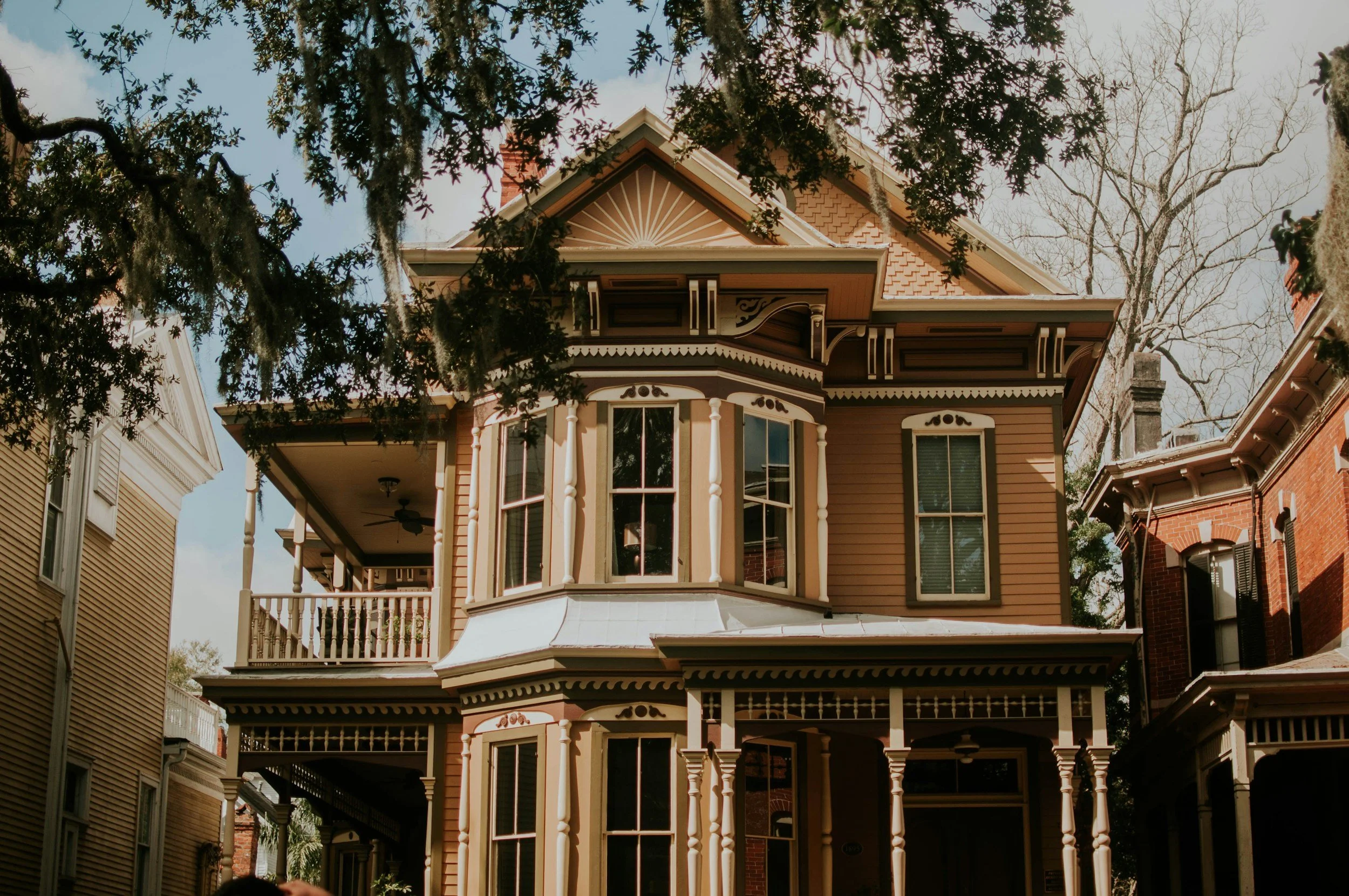 A detailed Victorian-style house with ornate woodwork, large windows, and a front porch, surrounded by trees, with a blue sky background.