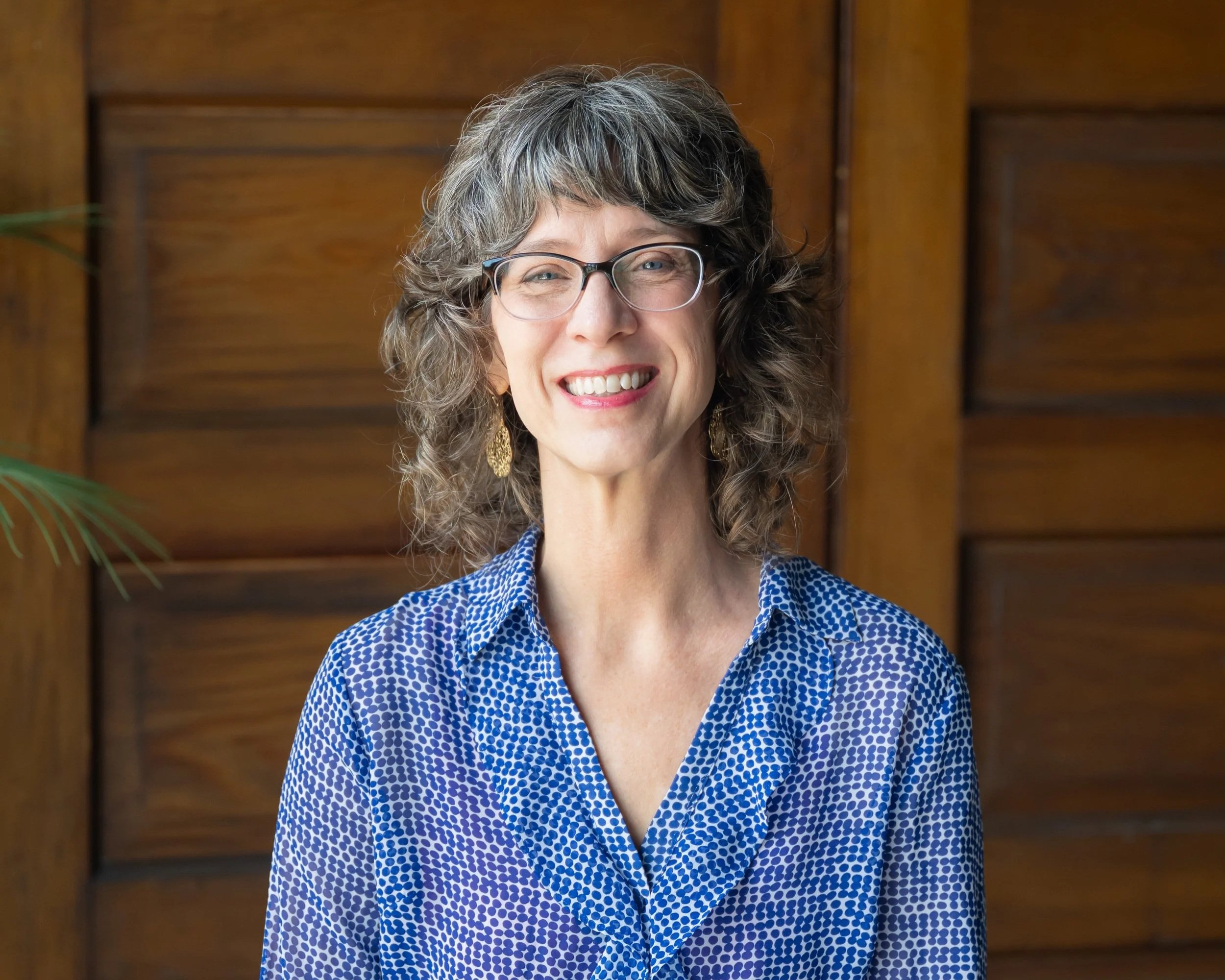 A woman with curly gray hair and glasses, smiling, wearing a blue patterned blouse and gold earrings, standing in front of a wooden background.
