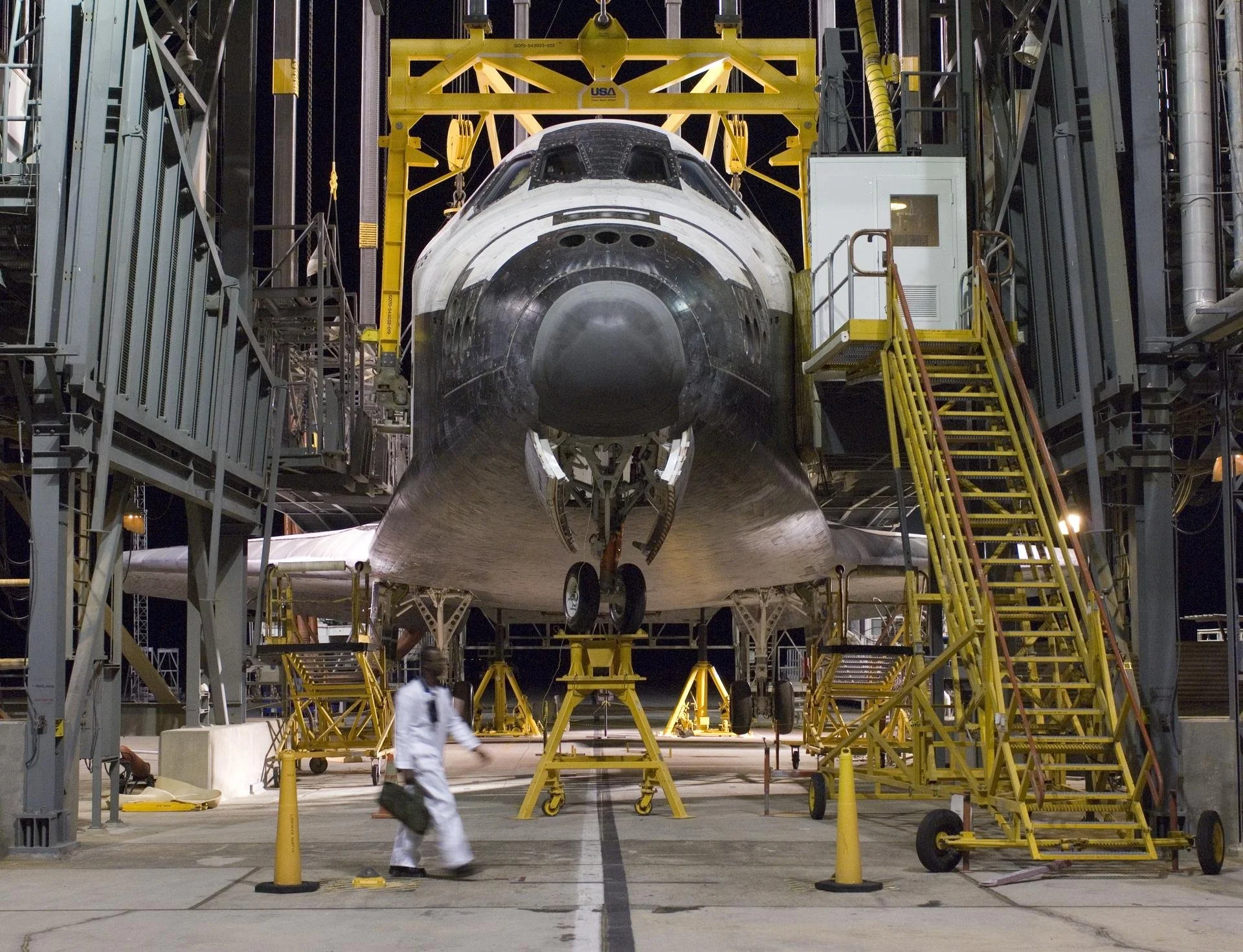 Inside a large space facility, a space shuttle is being assembled or maintained with scaffolding and equipment around it. The shuttle's nose is visible in the foreground, with a person in a white protective suit walking nearby.
