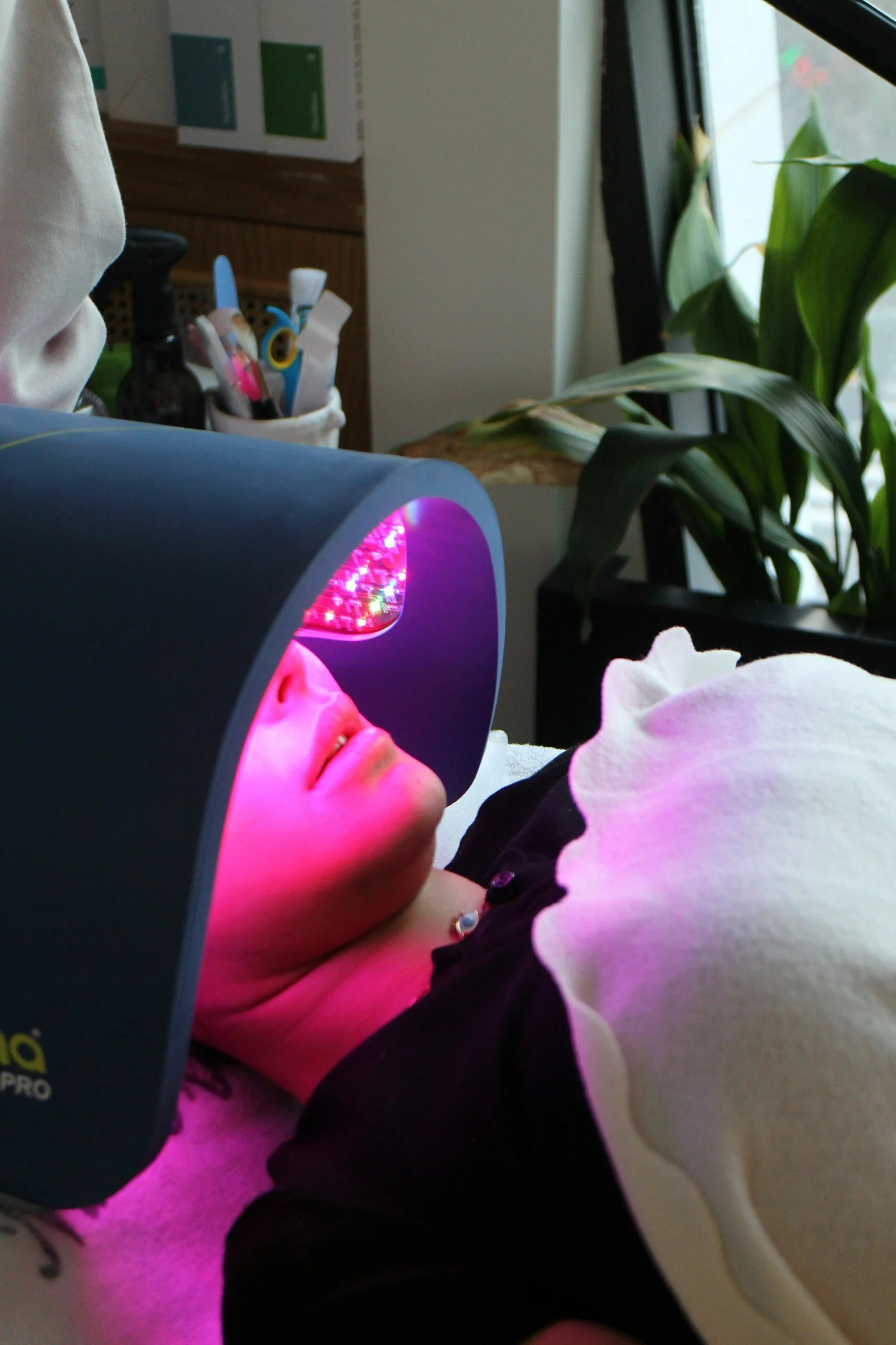 A young girl with glasses is lying down with her eyes closed, illuminated by purple-pink light from a device near her face. She is barefoot and wearing a black and white shirt, resting on a soft surface near greenery and household items.