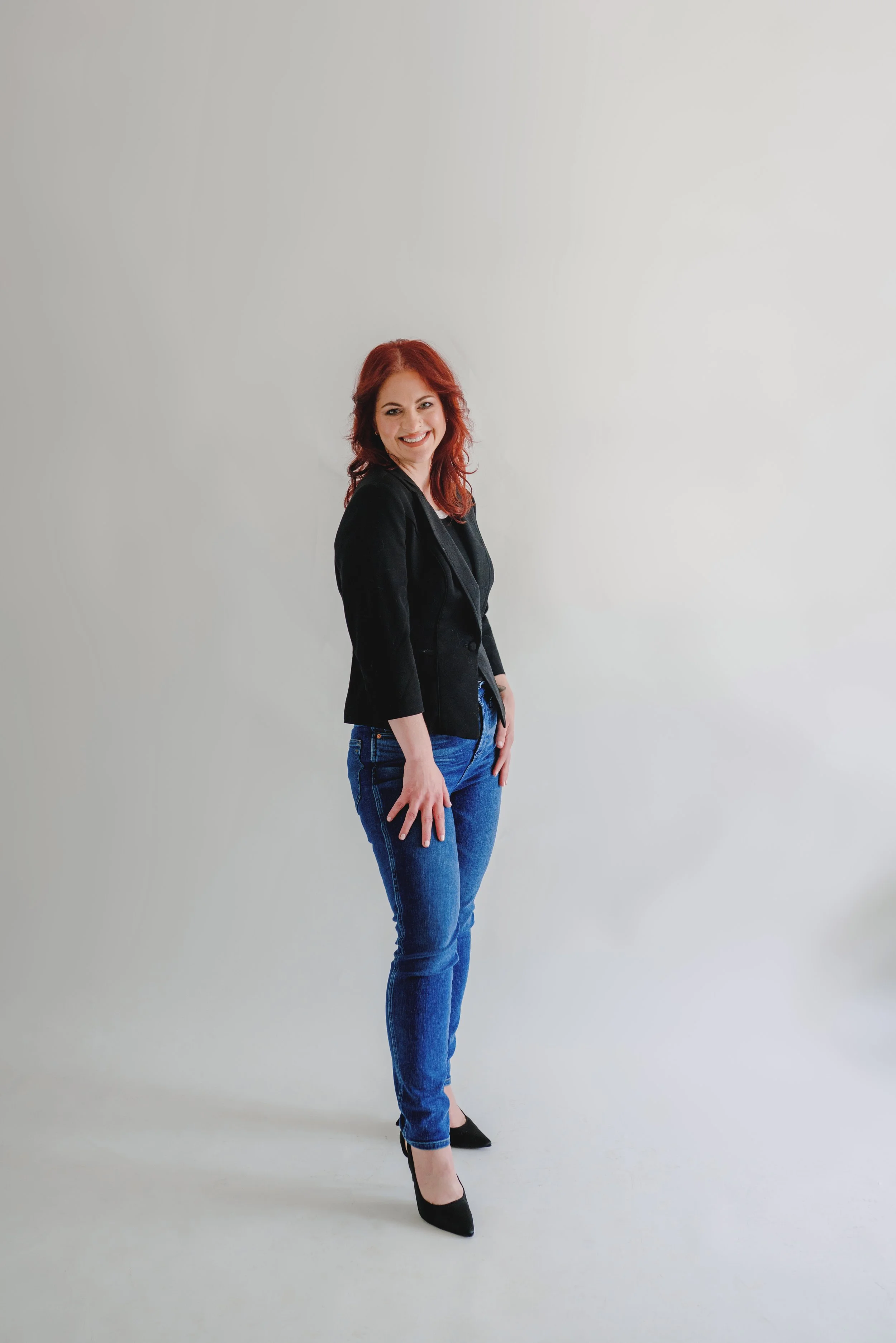 A woman with red hair wearing a black blazer, black heels, and blue jeans, standing against a plain white background, smiling and posing with one hand by her side.
