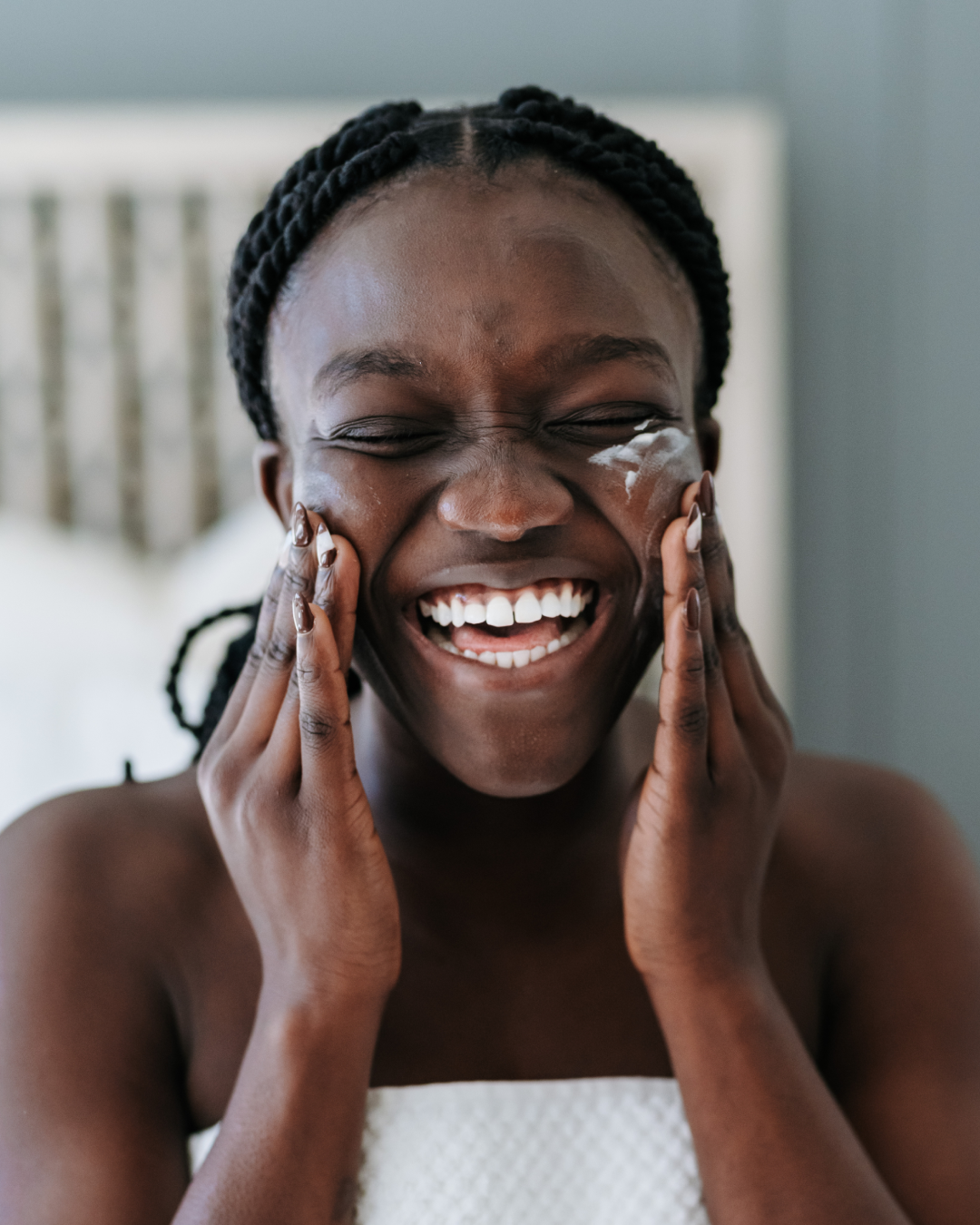 A woman with braided hair laughing and applying facial cream while sitting on a bed.