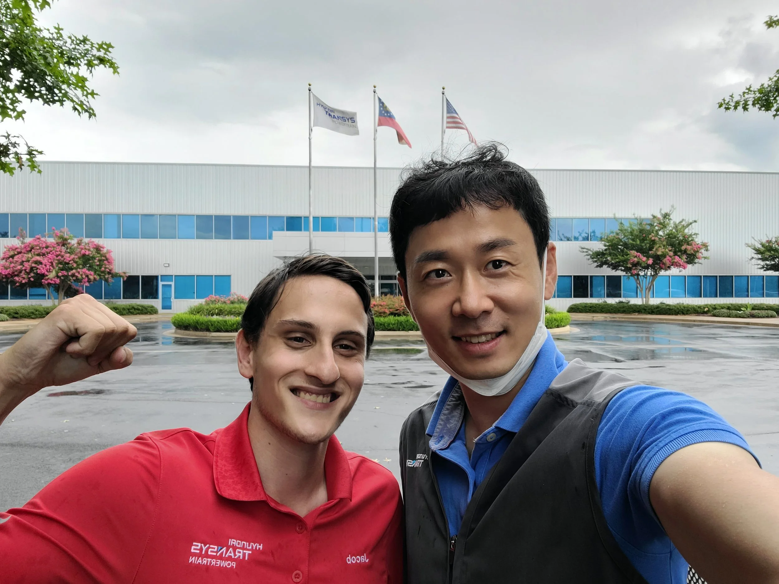 Two men are smiling and taking a selfie in front of a building with flags flying on flagpoles. The man on the left wears a red shirt and the man on the right wears a blue shirt. There are flowering trees and a cloudy sky.