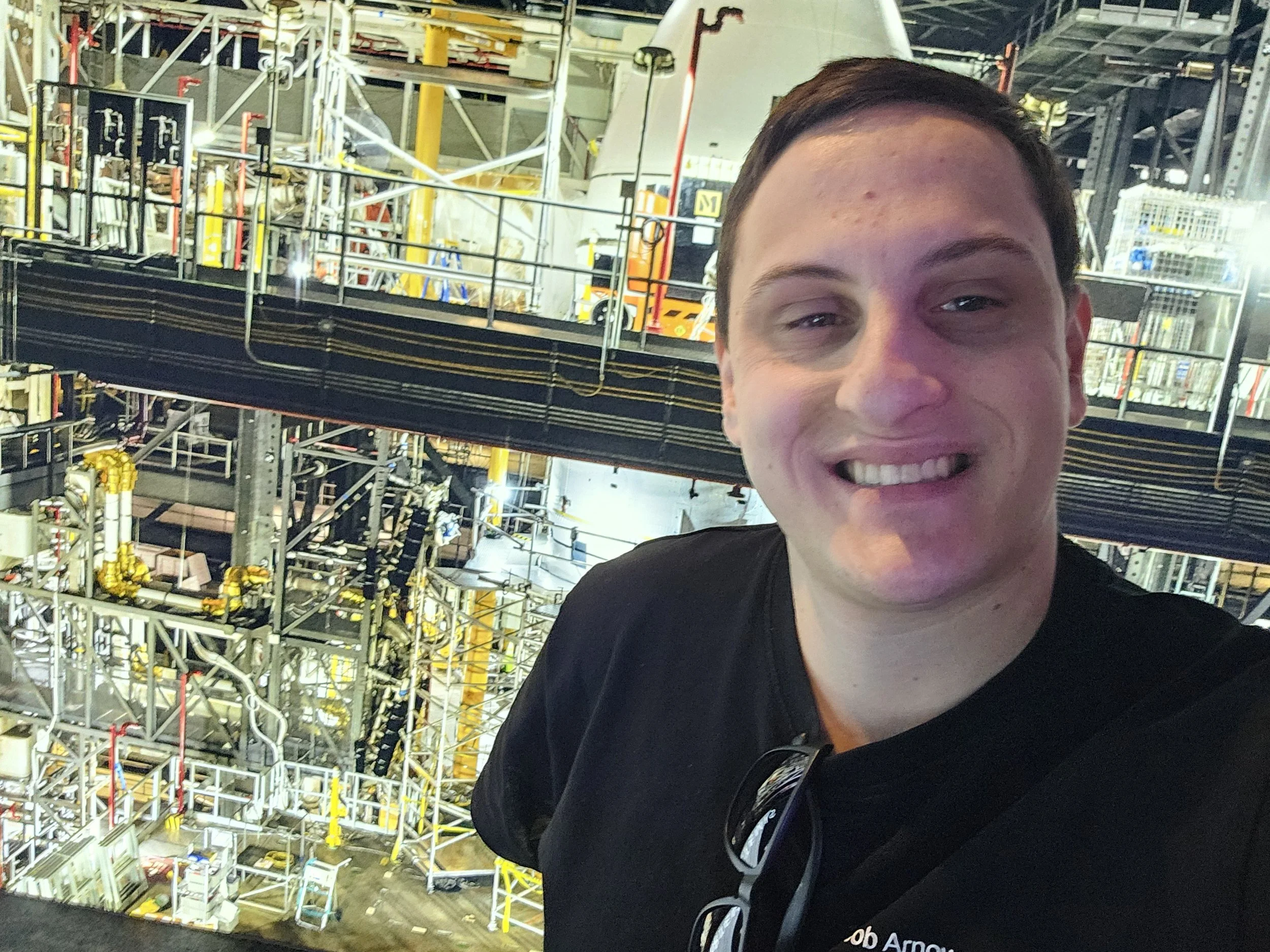 A person taking a selfie inside an industrial facility, possibly a power plant or factory, with complex machinery, pipes, and scaffolding in the background.