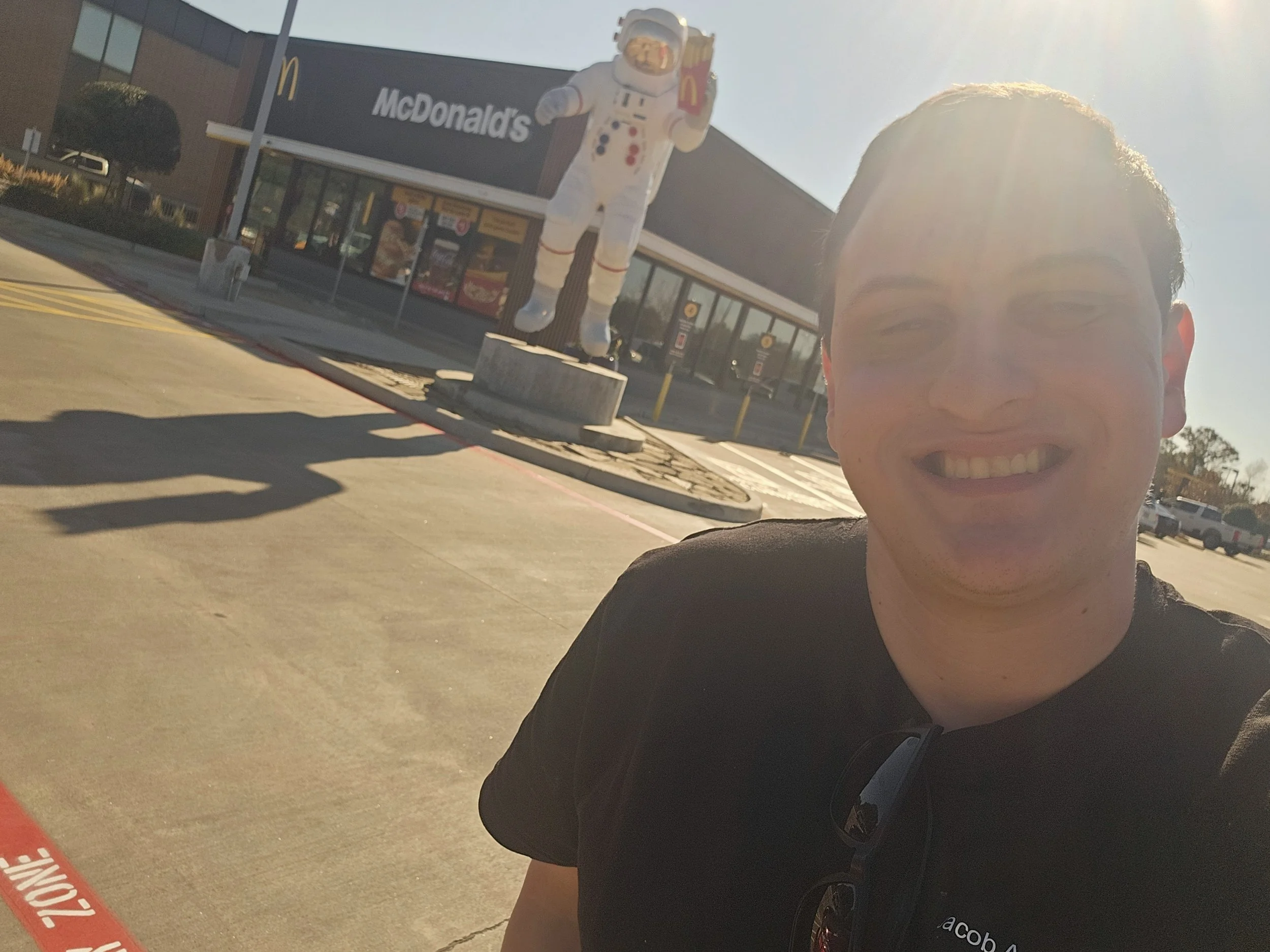 A young man taking a selfie in a parking lot with a McDonald's restaurant and a large astronaut figure in the background.