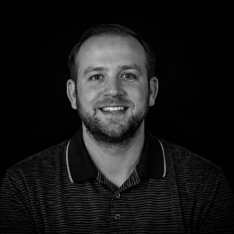 A black and white portrait of a smiling man with short hair and facial hair, wearing a striped collared shirt.