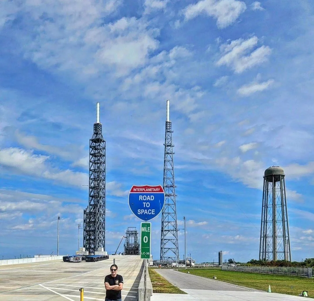 A person standing in front of launch pads and antennas at a space launch site with a sign that reads 'Road to Space,' indicating a space exploration theme.