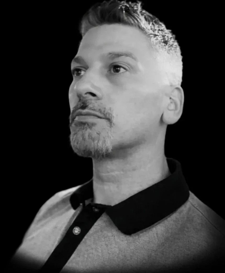 Black and white portrait of a man with short, styled hair and a goatee, wearing a polo shirt with a dark collar, against a dark background.