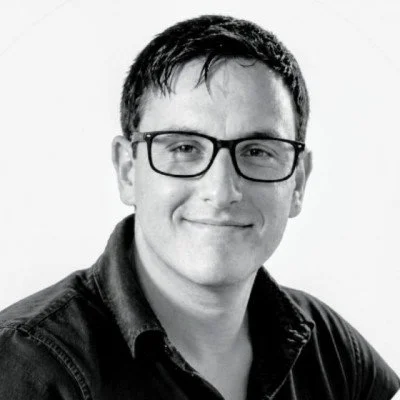 Black and white portrait of a young man wearing glasses and a dark shirt, smiling gently.