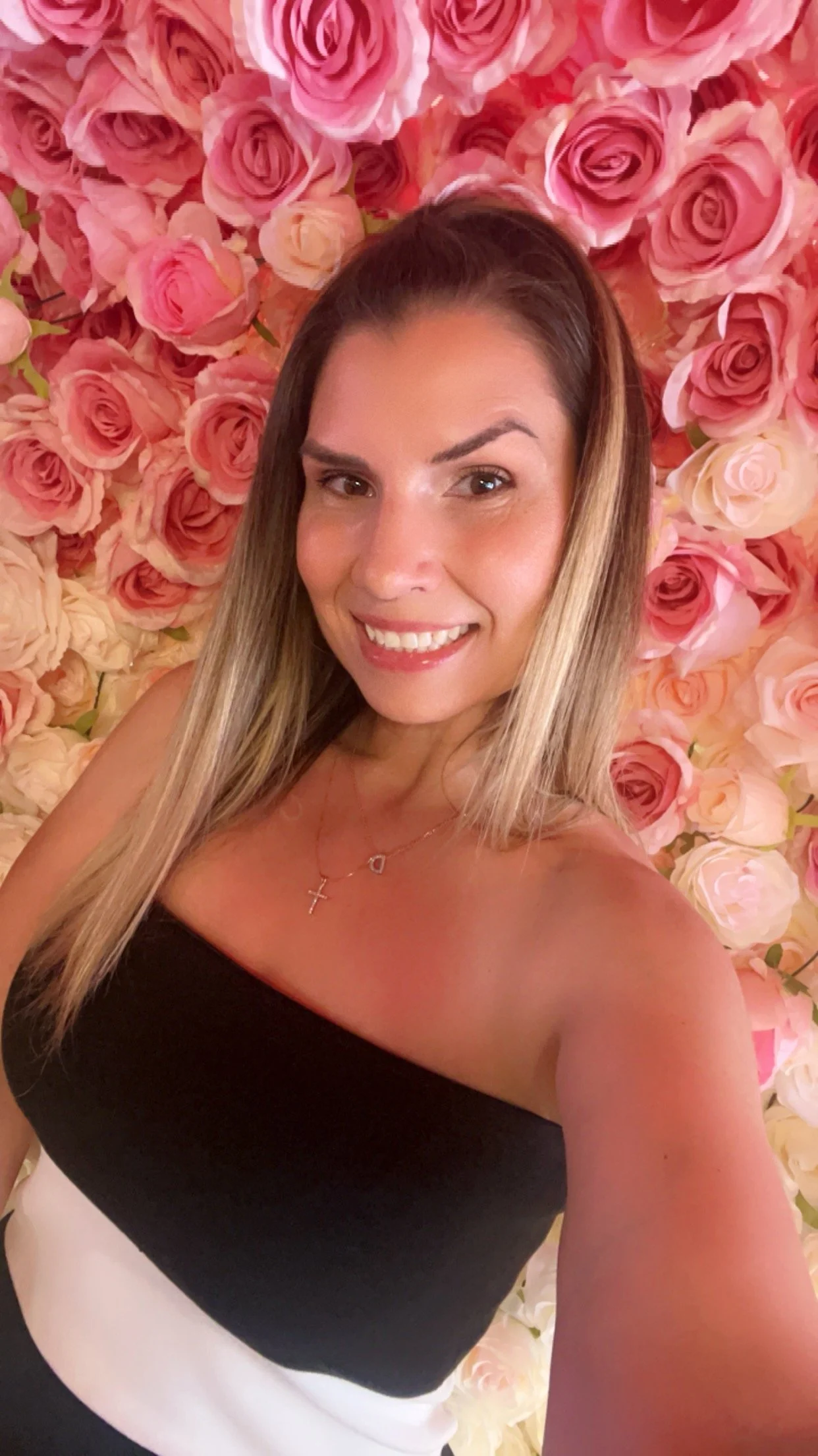 A woman smiling taking a selfie in front of a backdrop filled with pink and white roses.