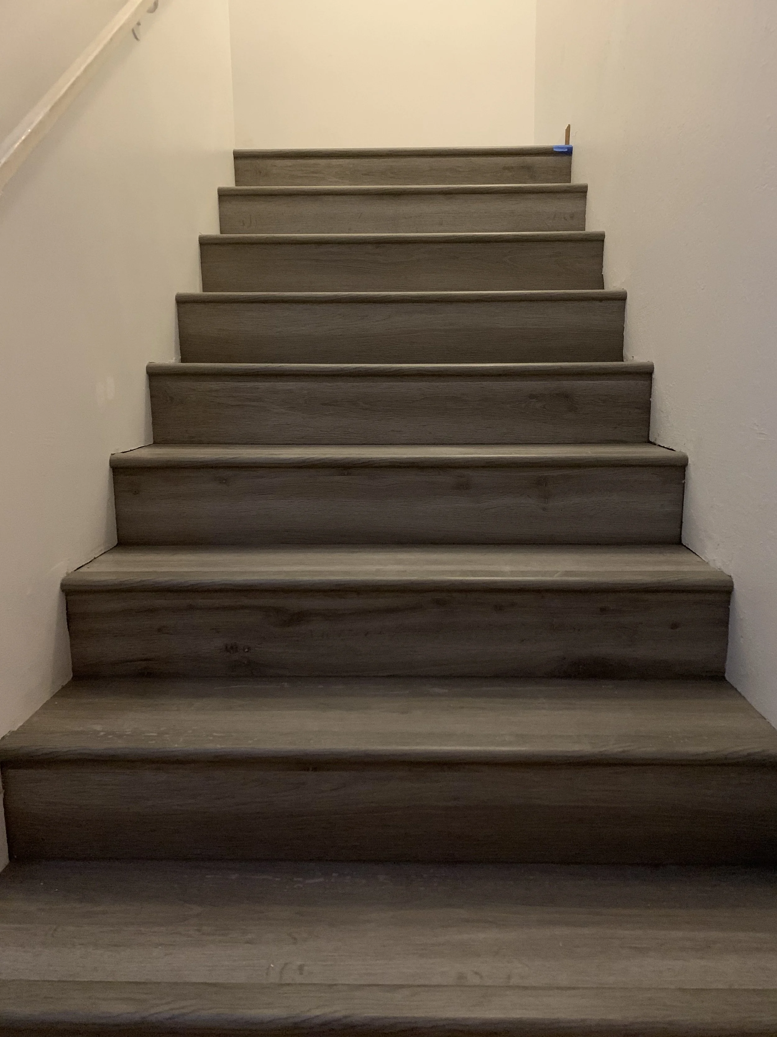 LVP staircase with eight steps, beige walls, and a small blue object or tape on the upper right corner of the top step.