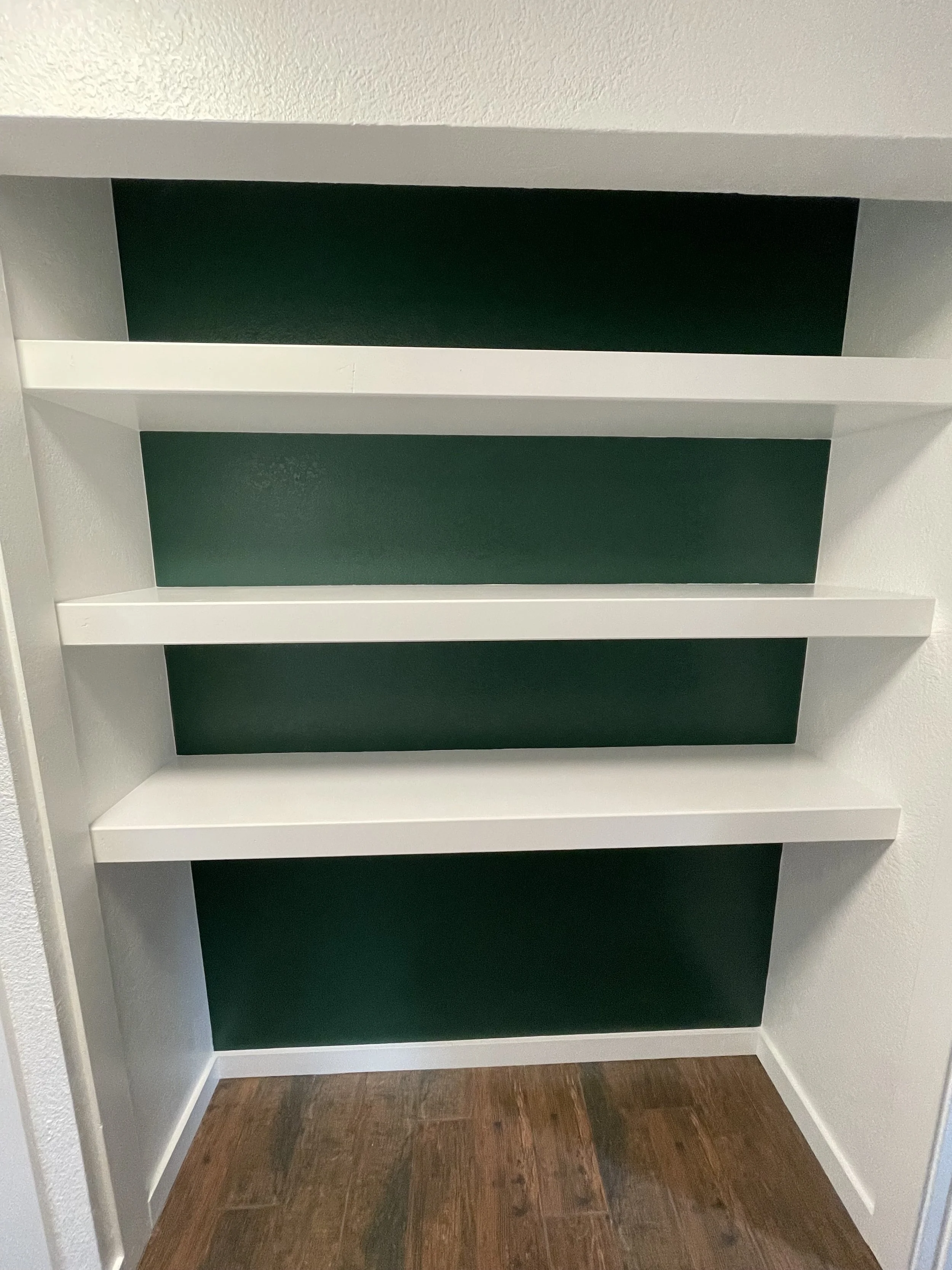 Empty custom white built-in shelves with a dark green back panel on a tile wooden floor.