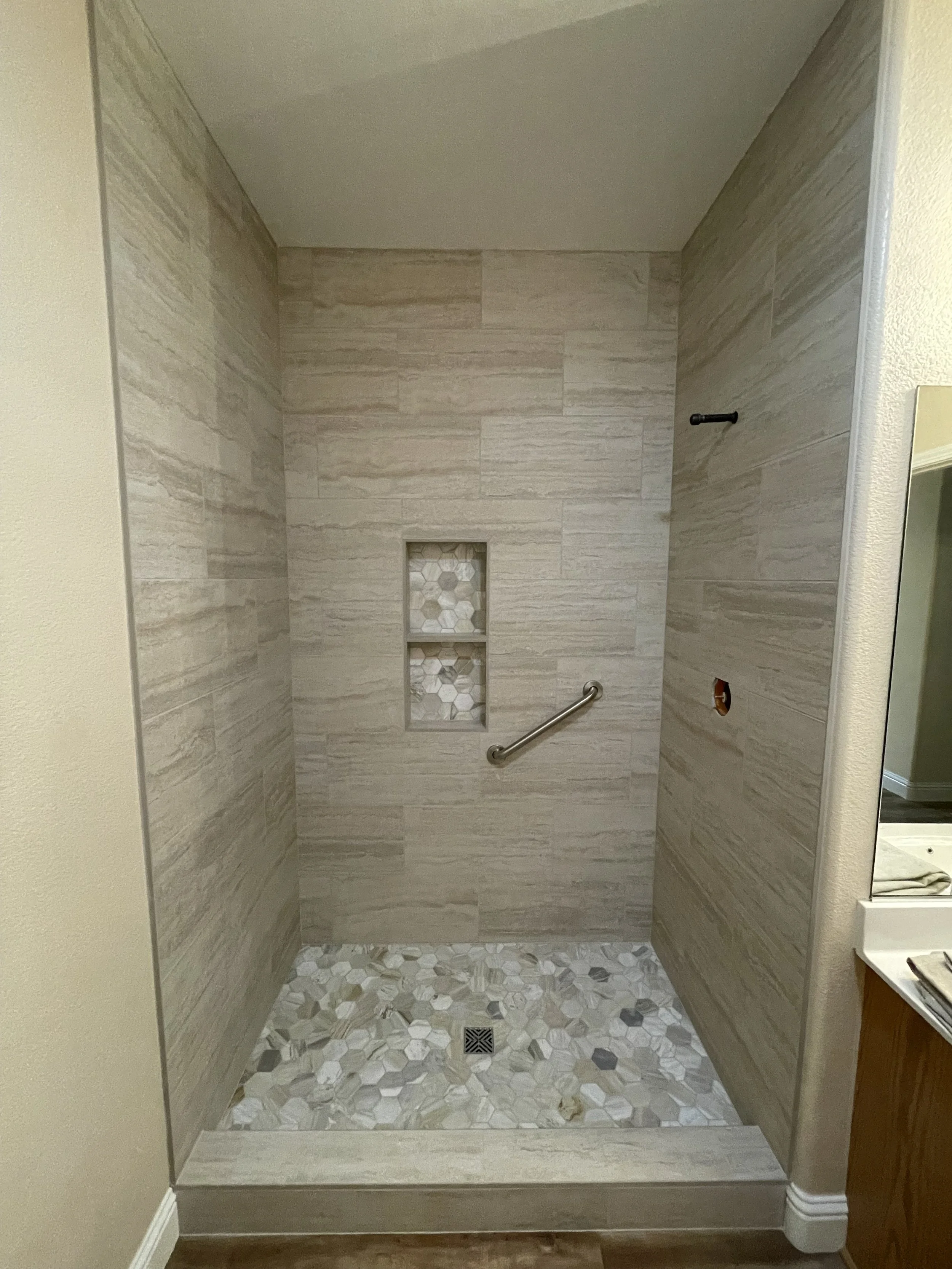 Empty shower with beige tile walls, hexagonal floor tiles, a small built-in shelf with mosaic tile, and a horizontal grab bar.