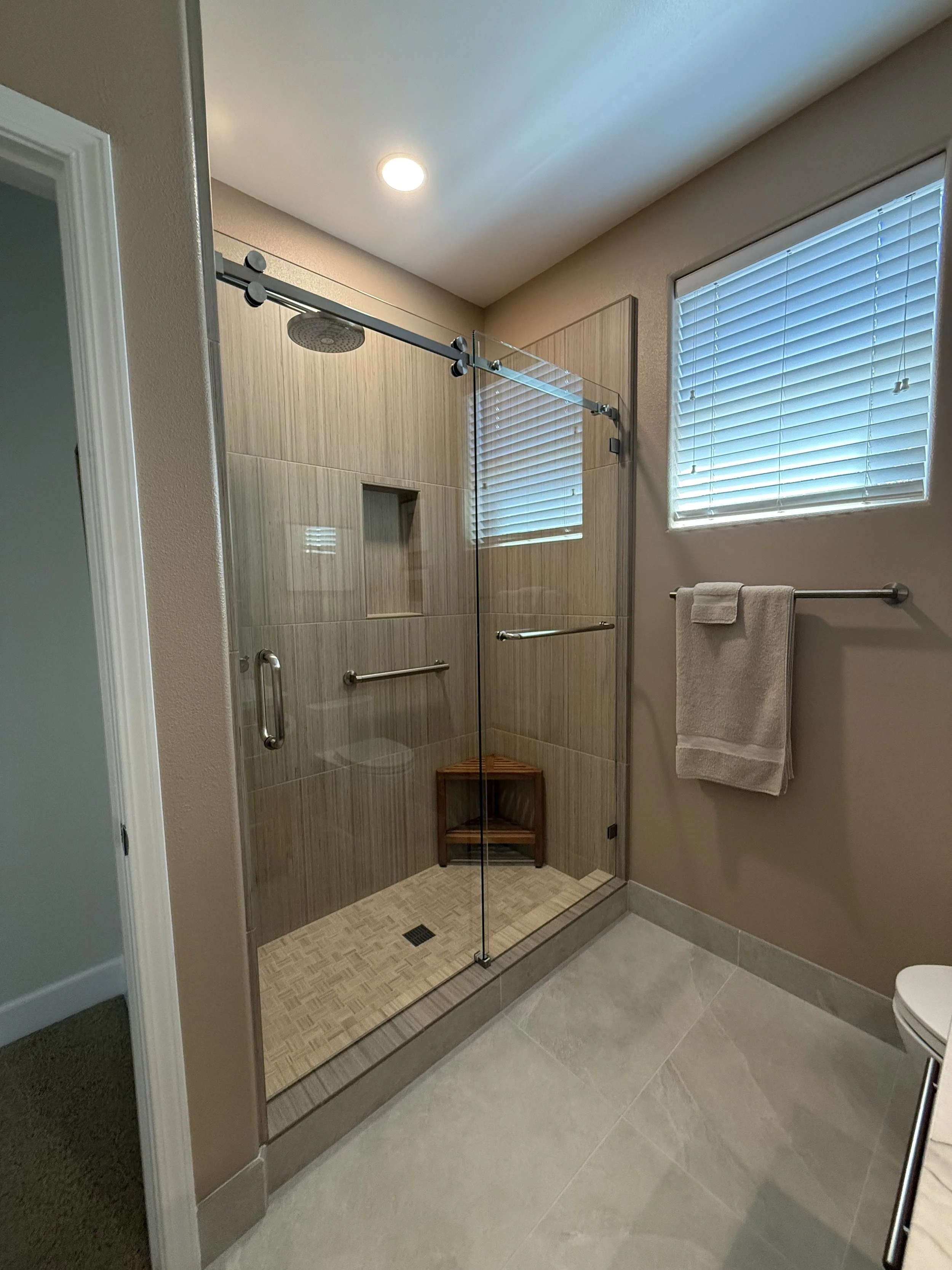 Modern bathroom shower with glass door, beige tiles, a built-in shelf, and a small wooden stool inside, with a window and towel rack on the adjacent wall.