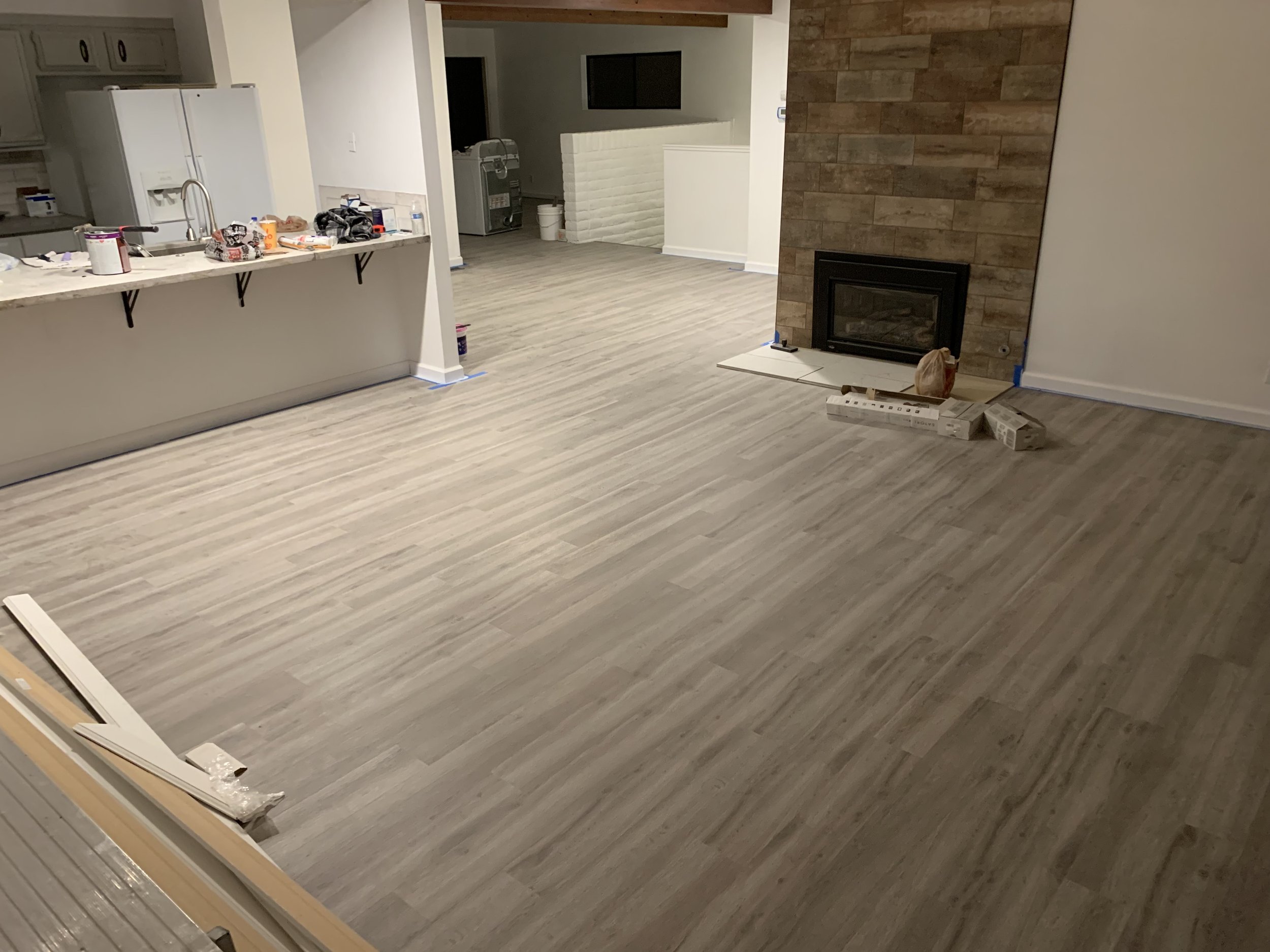 Living room with ongoing flooring installation, tools, and construction materials scattered around, featuring a fireplace with a wood-paneled wall.